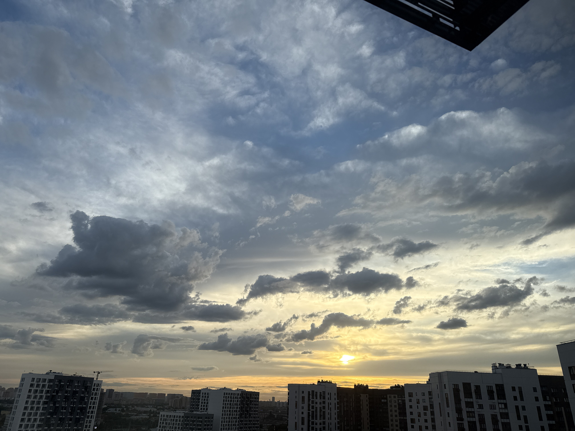The image captures a dynamic sky over the city of Lyubertsy, Russia, likely taken during sunset. Dominating the upper portion of the frame are dramatic, voluminous clouds in shades of gray, white, and light blue, suggesting a partly cloudy or overcast condition. The clouds are textured and varied, with some appearing wispy and others more dense. Near the horizon, the sky transitions to warmer tones of yellow and orange, indicating the sun is low in the sky and possibly partially obscured by clouds. A small, bright spot of sunlight is visible through a gap in the clouds.
In the foreground and midground, the rooftops of numerous modern apartment buildings are visible. These buildings are characterized by their geometric designs, with contrasting dark and light facades. Many of them have a significant number of windows and appear to be mid to high-rise structures. A construction crane is visible on the left side of the frame, suggesting ongoing development in the area. The overall impression is of an urban landscape under a dramatic and colorful sky. No people or specific actions are discernible in the image.