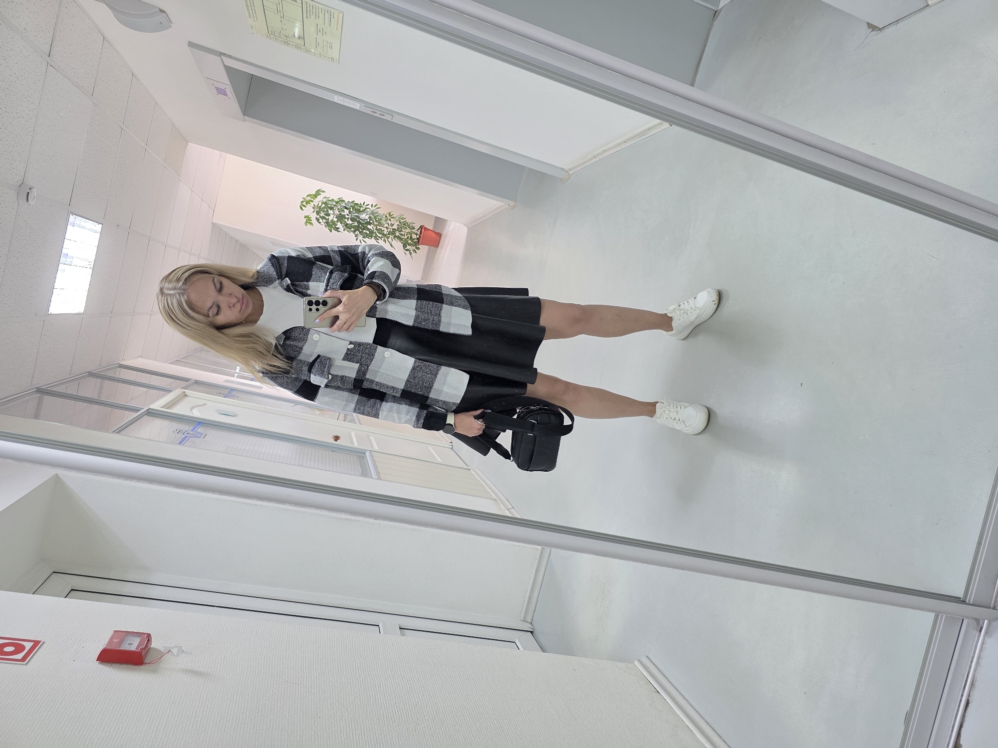 A young woman with blonde hair is standing in a hallway, taking a selfie in a large mirror. She is wearing a black and white checkered shirt over a white t-shirt, a black leather skirt, and white sneakers. She is holding a black handbag in her left hand and her smartphone in her right hand. The hallway appears to be in an office building or a similar institutional setting, with white walls, a tiled ceiling, and a grey floor. A red fire alarm box is visible on the left wall. There is a potted plant and an elevator door in the background. The image captures a moment of self-presentation and everyday life within a nondescript urban environment.