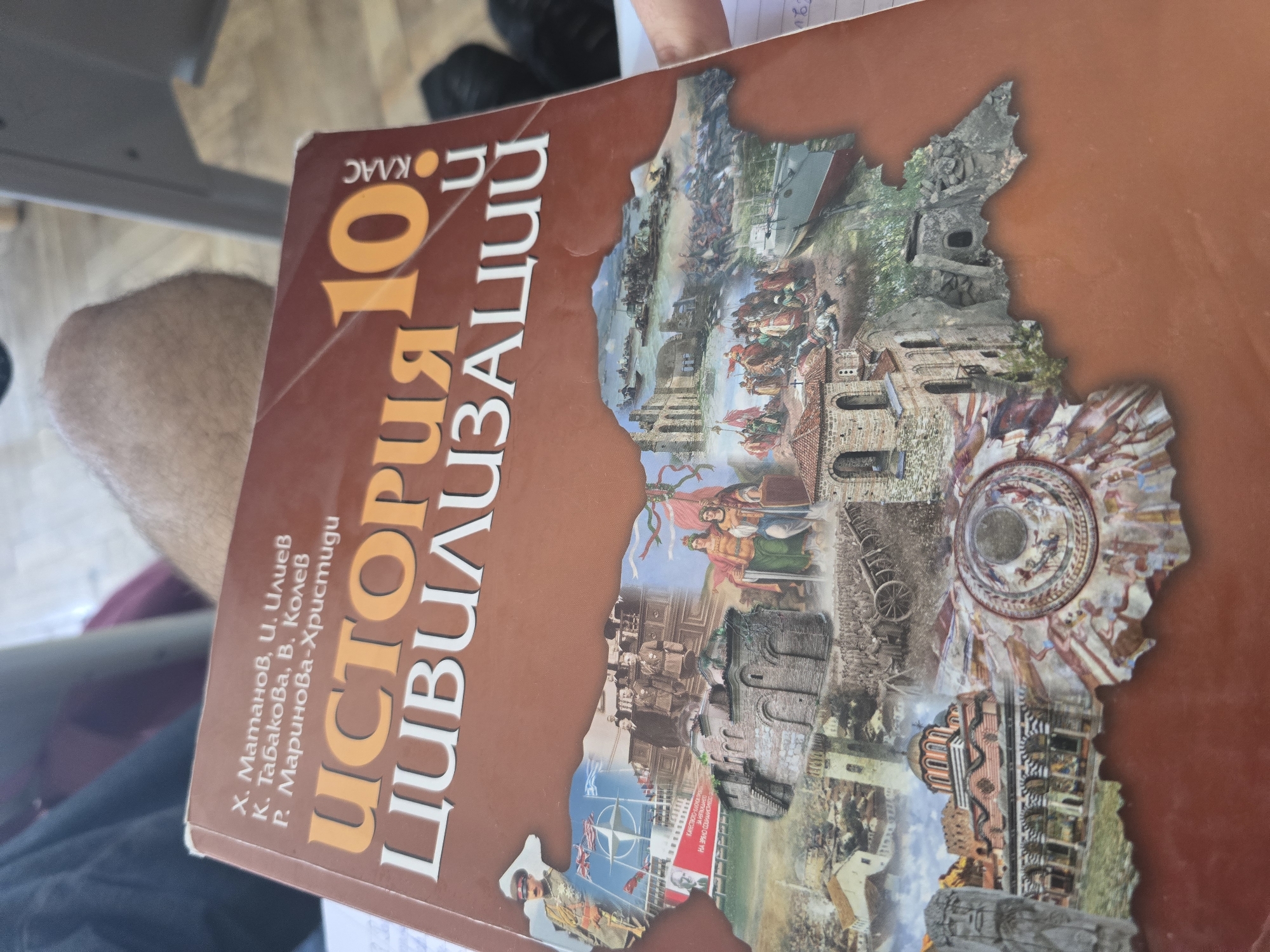The image displays a Bulgarian textbook titled "ИСТОРИЯ 10 КЛАС ЦИВИЛИЗАЦИИ" (HISTORY 10th Grade CIVILIZATIONS), held at an angle in what appears to be an indoor setting in Sofia, Bulgaria. The textbook's cover is predominantly brown and features a rich collage of historical imagery, arranged within an outline that strongly resembles the map of Bulgaria.
The detailed collage on the cover showcases various historical periods and cultural elements relevant to the study of civilizations. Visible illustrations include dynamic battle scenes with cavalry and foot soldiers, architectural fragments of ancient or medieval buildings, distinct Orthodox churches with frescoes and domes, a cannon, a sailing ship, and portraits of historical figures. More modern elements are also present, such as the NATO flag and a red banner containing text that reads "КАЧЕСТВОТО-ТАЛЮНО НА ЗРЯЛО СОШИАЛИСТИ" (QUALITY - THE TALENT OF MATURE SOCIALISTS), hinting at Bulgaria's socialist past. The number "100.00" is also visible on this banner.
The authors' names are listed on the left side of the cover: Х. Матанов, И. Илиев, К. Табакова, В. Колев, and Р. Маринова-Христиди. The text "10 КЛАС" clearly indicates that the book is intended for 10th-grade students. The background is blurred, showing a person's arm or leg and a light-colored surface, possibly a floor or desk, consistent with a classroom or study environment.