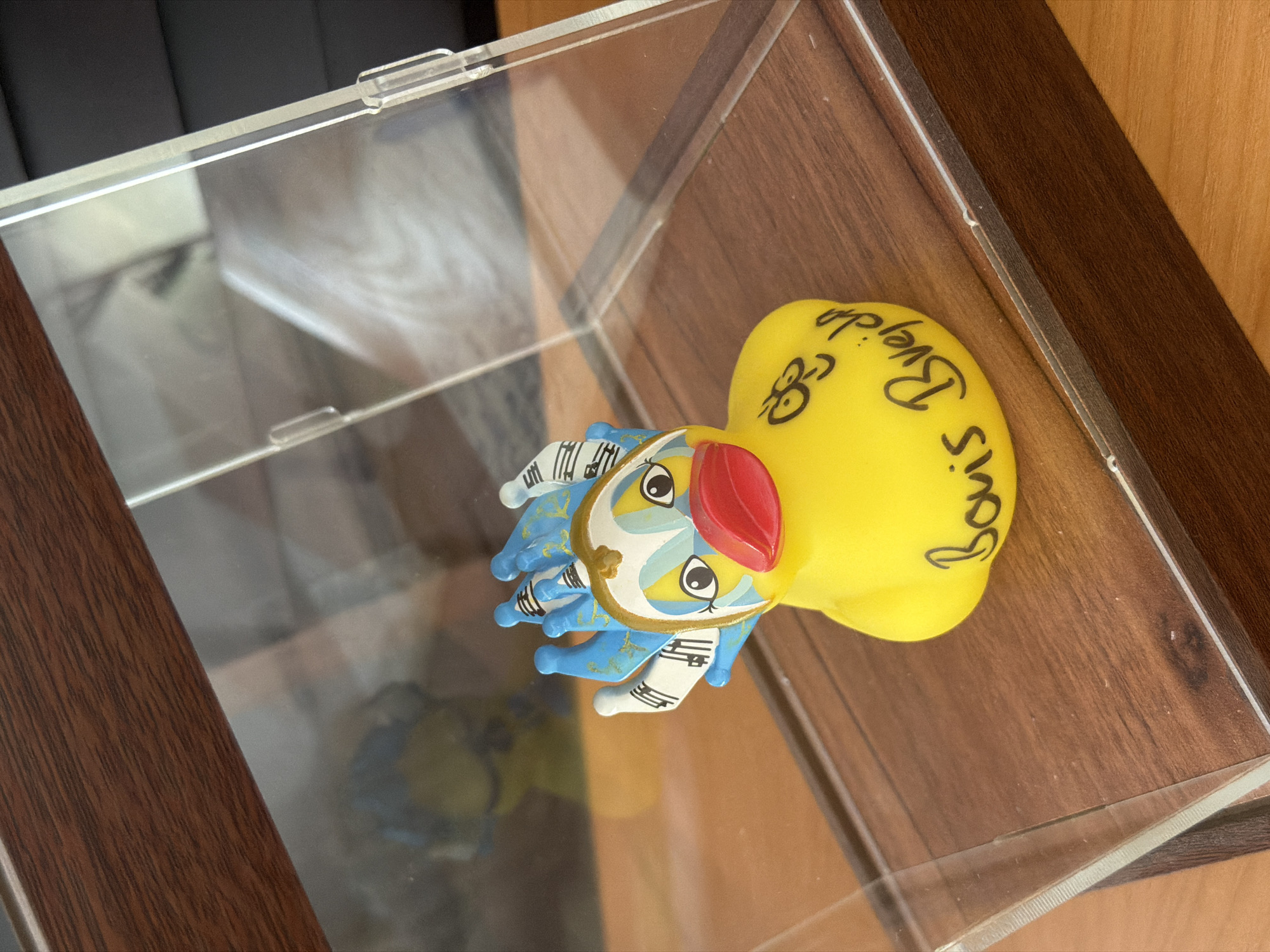 A yellow decorative rubber duck with a painted face and blue and white headpiece is displayed inside a clear plastic case. The duck has large, cartoonish eyes and a bright red beak. The headpiece is decorated with musical notes and has several blue points. Black cursive writing on the duck's body reads "Boris Brejcha." The duck sits on a wooden surface within the case, which is situated on a larger wooden surface. The background is blurred and appears to be indoors. The exact location in Baan Plai Laem, Thailand, is not discernible from the image.