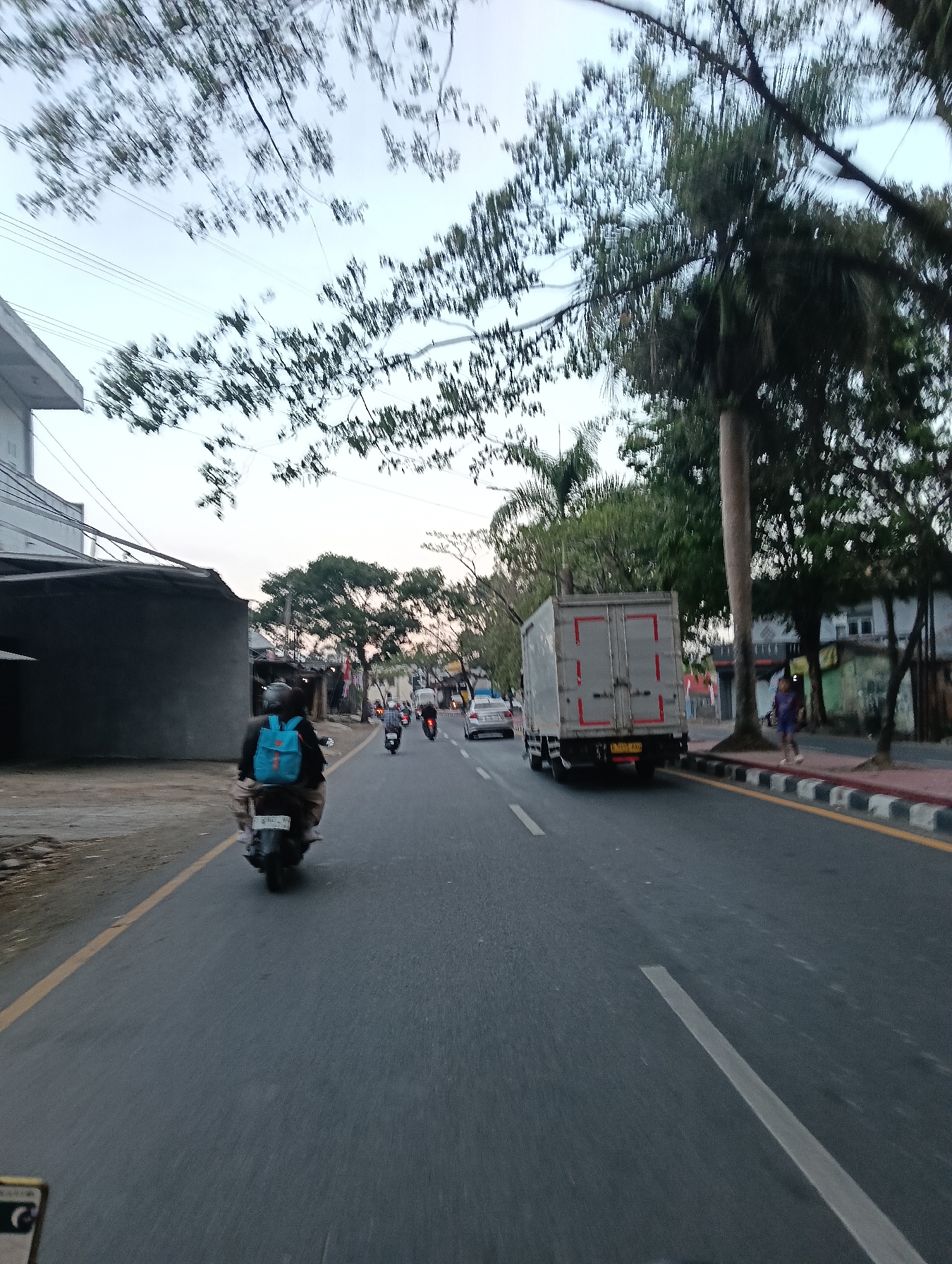 The image captures a street scene in Tasikmalaya, Indonesia. In the foreground, a person is riding a motorcycle, distinguished by a bright blue backpack. The road is paved and marked with yellow and white lines. Traffic is present, with a white box truck and other vehicles visible on the road. Trees line the street, and buildings are situated on the right side. The sky appears overcast. The overall impression is that of a typical day in a bustling urban environment in Indonesia, likely during the late afternoon or early evening given the lighting.