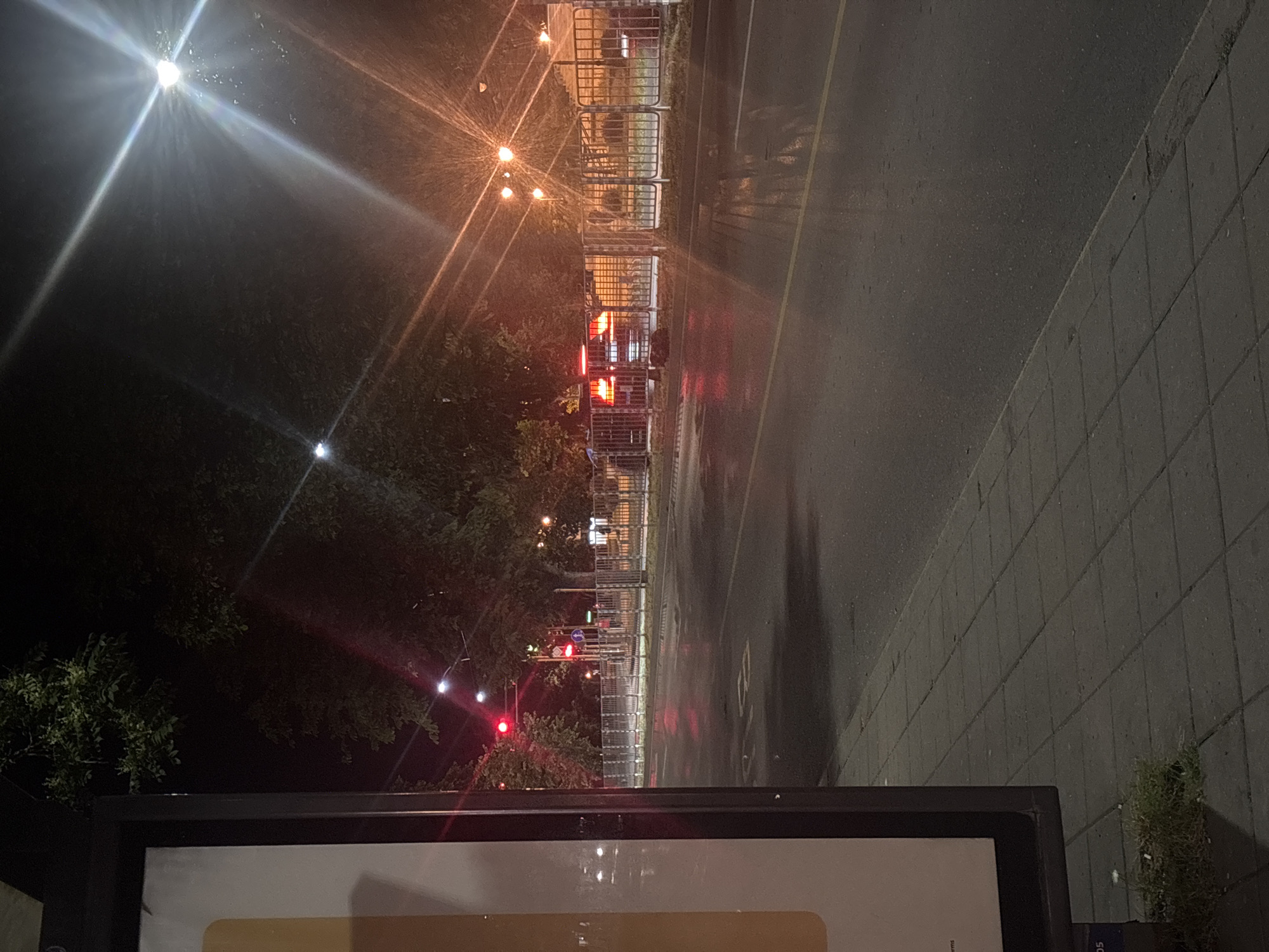 The image captures a nighttime scene on a city street in Varna, Bulgaria. A traffic light displaying a red signal is visible, indicating that vehicles should stop. Several cars are present, some with their brake lights illuminated, suggesting they are stopped at the intersection. The street appears wet, reflecting the lights from the traffic signals and street lamps, creating a shimmering effect. Trees line the side of the road, their branches dark against the night sky. In the foreground, a sidewalk made of concrete tiles is visible, and to the left, part of a large advertising billboard is in frame. The overall atmosphere is calm and quiet, characteristic of a city at night.