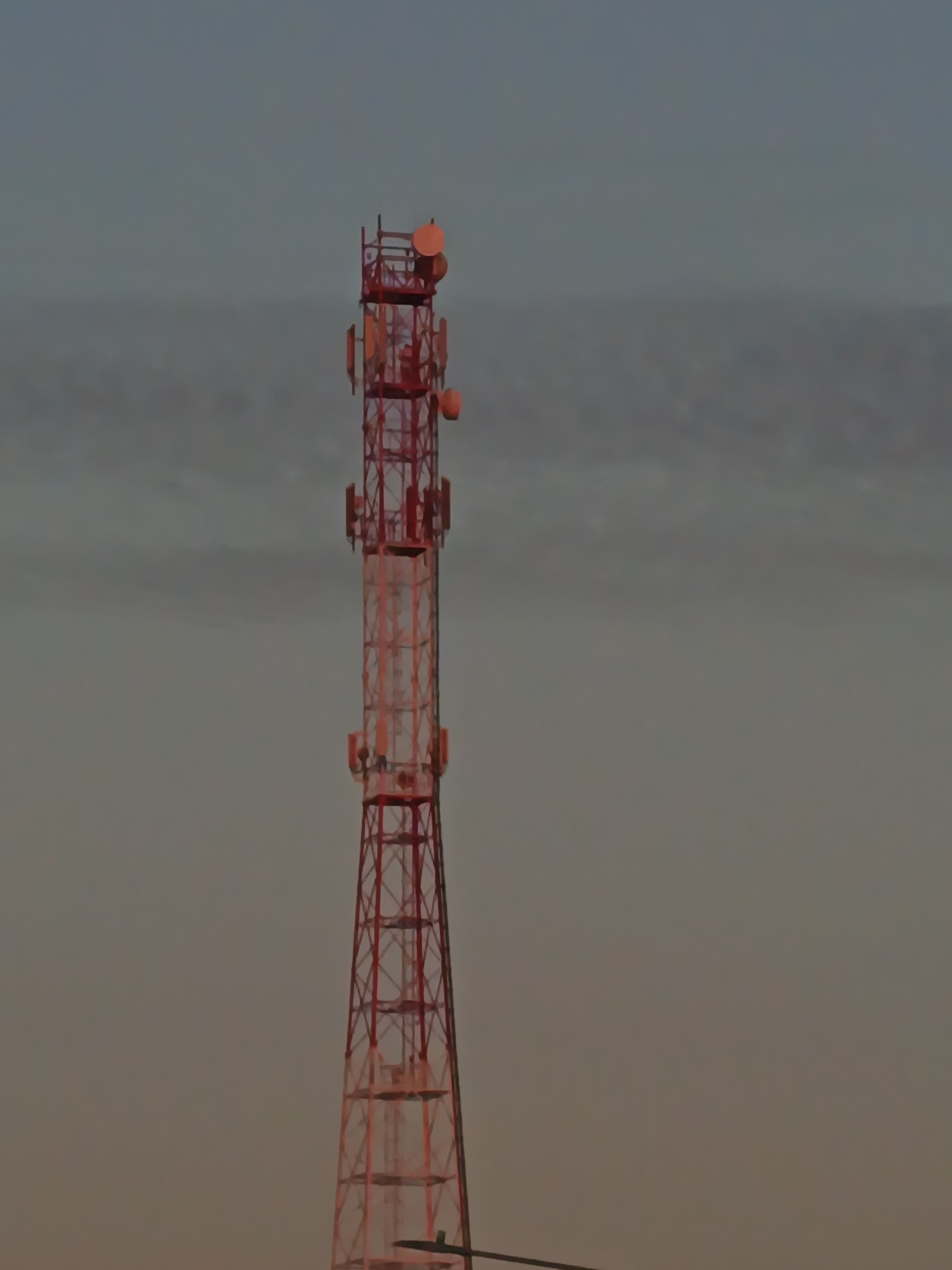 An outdoor, medium shot captures a tall, red lattice cell tower against a muted, cloudy sky. The tower is centrally framed and its metallic structure is visible, with various antennas and dishes attached at different levels. The sky displays a gradient of soft grays and blues, suggesting either dawn or dusk. The overall impression is one of an industrial landscape in Воскресенское, Russia.