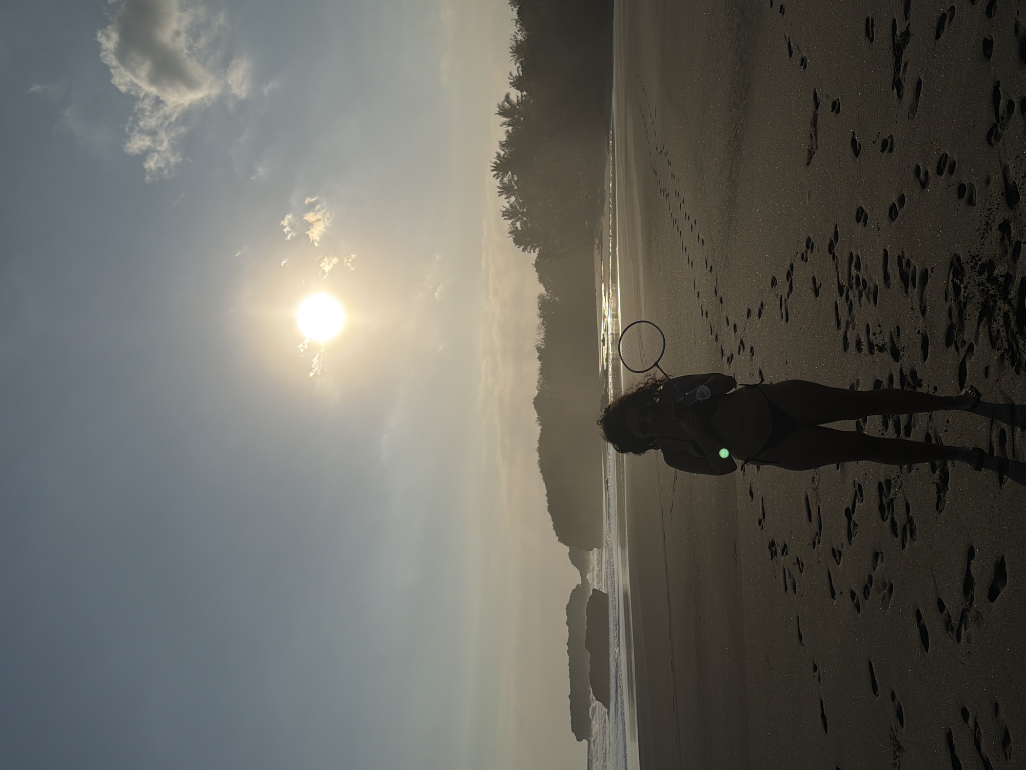 The image captures a backlit beach scene in Selemadeg, Indonesia, during late afternoon or early evening. The sky, a gradient of pale blue to a bright warm glow near the horizon, features a prominent, low-angle sun in the upper left quadrant, causing a strong silhouette effect and a green lens flare near the subject's midsection. Scattered cumulus clouds are visible in the upper sky. The ocean on the left exhibits moderate breaking waves near a dark sand beach.
Along the coastline on the left, several large, dark rock formations are partially submerged in the water. On the right, a dense line of tropical vegetation, primarily palm trees, borders the beach, appearing as a dark silhouette against the hazy light. The sandy beach in the foreground and mid-ground is dark, likely volcanic, and subtly glitters in the sunlight. Numerous human footprints are embedded in the sand, traversing various directions across the frame.
A single individual, appearing female, stands centrally in the lower half of the image, facing slightly to the right of the viewer's perspective. The person is presented in full silhouette, obscuring facial features, and has curly hair. They are wearing a dark two-piece swimsuit. In their left hand, they hold a badminton racket, and in their right hand, they hold a clear plastic bottle. The individual's bare feet are visible on the sand.