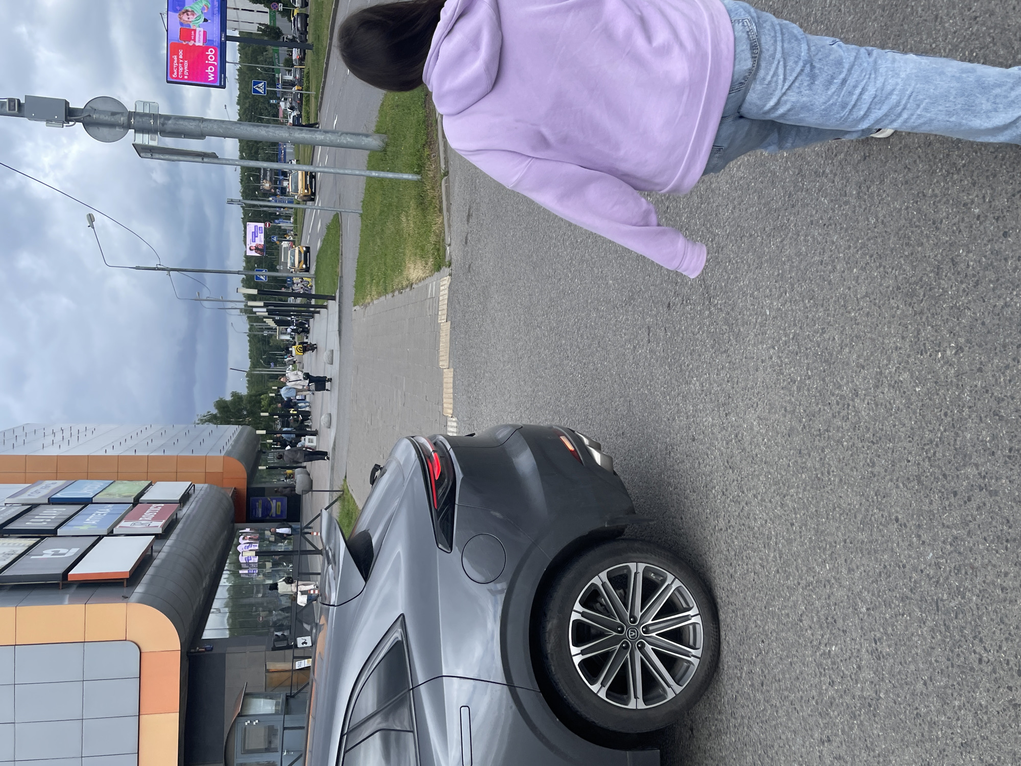 A young woman with dark hair, wearing a light purple hoodie and blue jeans, is walking away from the camera on an asphalt road. She is in the foreground, with the rear of a dark gray car on her left. The car has a distinctive wheel design.
In the background, there is a large building with an orange and gray facade, likely a shopping center, with various signs visible on its side. Beyond the building, a street lined with trees and streetlights is visible. Several cars and people are scattered along the street, suggesting it is a public area. There are also billboards along the street, one of which displays the text "wb job" and some Russian text.
The sky is overcast with dark clouds, indicating a cloudy day. The overall scene appears to be a commercial or urban environment, likely during daylight hours. The woman's posture and the context suggest she is simply walking through the area.