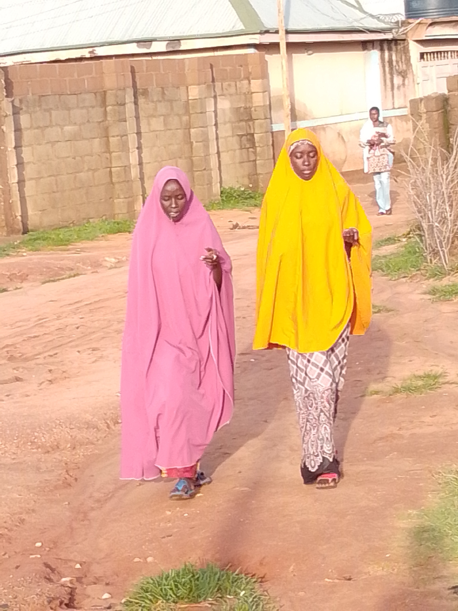 Two women walk on a dirt path in Gombe, Nigeria. The woman on the left wears a pink hijab and a long pink garment. The woman on the right wears a bright yellow hijab and a patterned skirt. Both women are wearing sandals. In the background, a man in a white and blue garment stands near a building. A brick wall borders the left side of the path, and sparse vegetation is visible on the right. The sun casts shadows, indicating it is likely late afternoon.