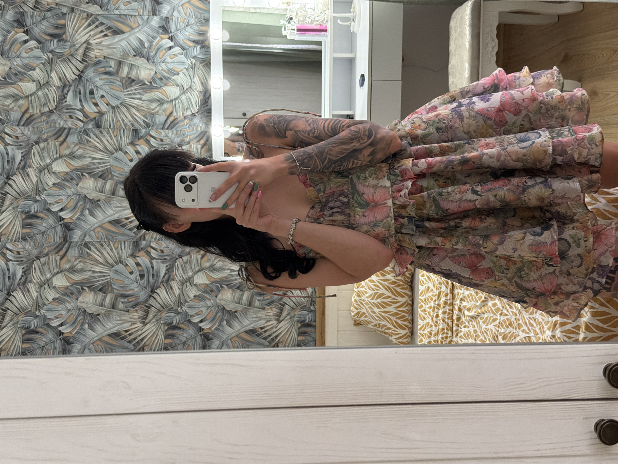 A person with long dark hair is seen taking a mirror selfie indoors, likely in a bedroom or dressing room in Plovdiv, Bulgaria. The individual is holding a white iPhone with three cameras, which obscures most of their face. They are wearing a sleeveless dress adorned with a vibrant floral and butterfly pattern, featuring shades of pink, green, purple, and beige. Extensive tattoos cover their left arm, and a delicate silver bracelet is visible on their left wrist. Their nails feature a green and pink/white patterned manicure.
The background, reflected in the mirror, displays a wall covered in a tropical leaf-patterned wallpaper in tones of grey, light blue, and beige. A large white vanity mirror with illuminated bulbs around its frame is visible, reflecting shelves containing various items. In the lower right of the reflection, a yellow and white patterned fabric, possibly bedding, can be seen. The foreground shows the white wooden frame of the mirror, characterized by horizontal planks and two dark, round drawer pulls at the bottom right. The word "iPhone" is faintly visible on the back of the smartphone. The indoor setting, with vanity lights on, suggests no specific time of day.