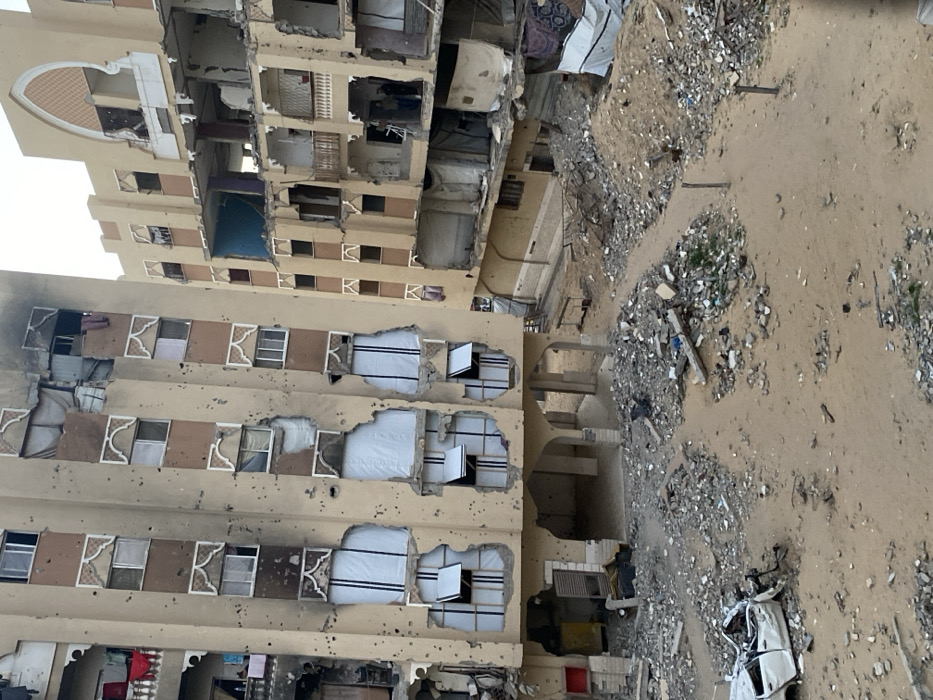 The image depicts a scene of extensive urban destruction in Khan Yunis, Palestinian Territory, under an overcast sky. Two multi-story buildings, consistent with residential structures, are the primary subjects, both exhibiting severe damage.
The building on the left, an approximately ten-story structure with a tan facade and ornamental arch-shaped window surrounds, is pockmarked with numerous small impact points, likely from bullets or shrapnel. Multiple windows are shattered or missing, and several sections of its exterior wall are blown out, revealing interior rooms. Some lower-story windows are covered with white, vertically striped material. Clothes hang on balconies on the upper left side of this building. The ground floor features arched open spaces.
The building on the right, also multi-story and tan, appears partially collapsed or significantly structurally compromised. Large portions of its exterior walls are missing, exposing multiple floor levels and interior rooms, some showing blue and purple paint. Its upper section features a large, decorative arched recess. On a lower-mid floor of this building, two indistinct human figures are visible standing within a damaged room, near an exposed edge.
The ground area between and in front of the buildings is covered in sand and extensive debris, including piles of concrete rubble, twisted metal, and miscellaneous refuse. A heavily damaged, overturned white vehicle, appearing to be a car, is situated in the lower-left foreground amidst the debris. Dirt tracks are visible on the sandy ground, weaving through the scattered rubble.