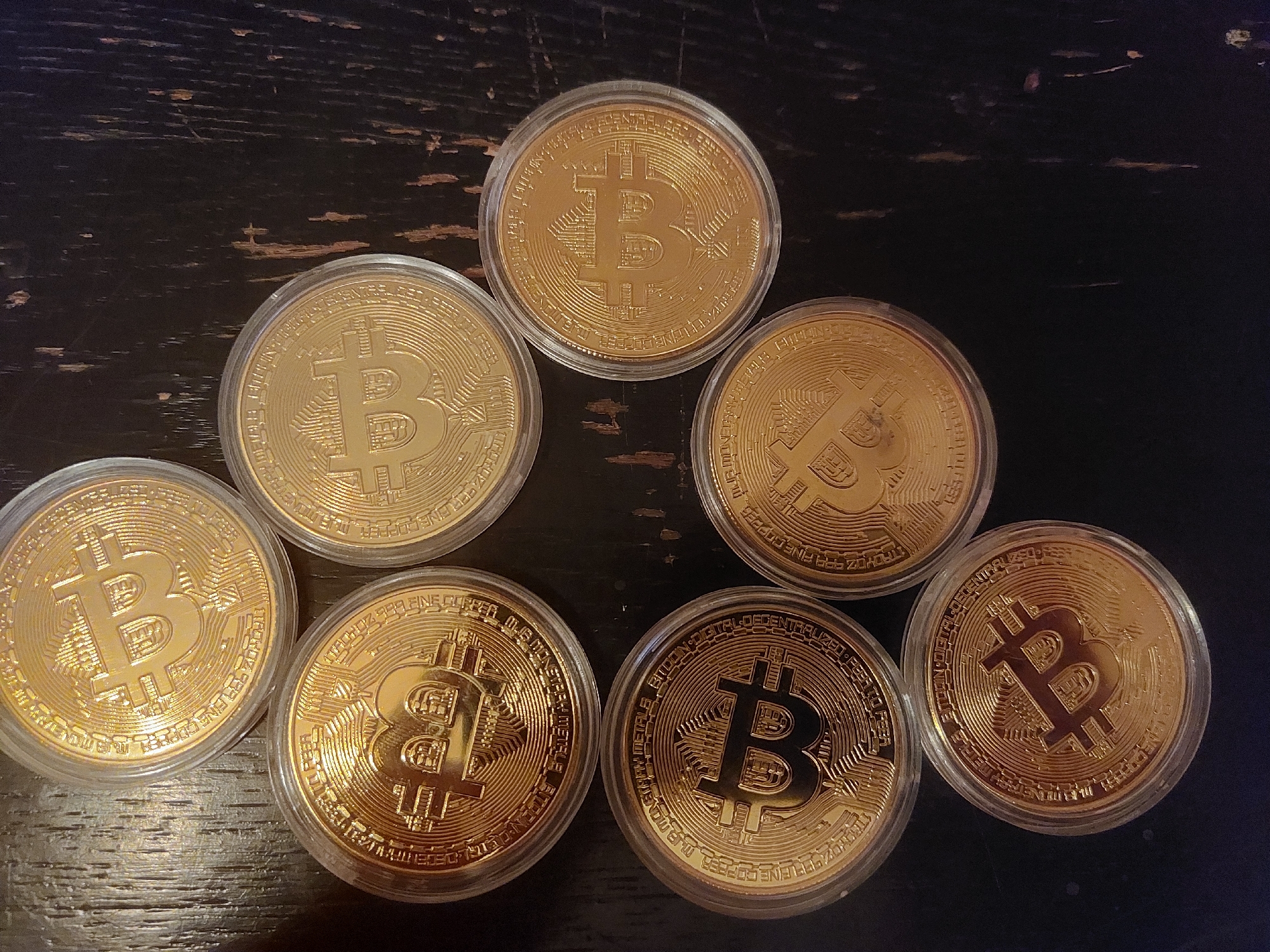 Seven golden Bitcoin coins are arranged on a dark wooden surface. The coins are housed in clear plastic cases. The coins are arranged in a semi-circular fashion. The background is a dark, possibly wooden, surface. There is no indication of the location being Tsarevo, Bulgaria.