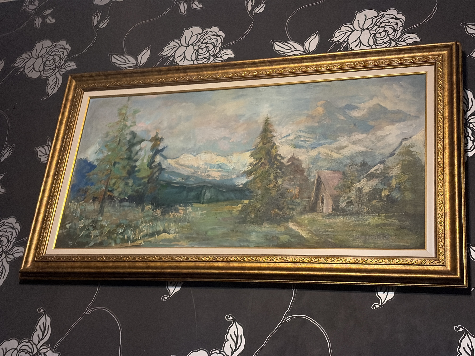 A framed landscape painting hangs on a wall covered in black wallpaper with a white floral pattern. The painting depicts a mountain scene with trees, a small building, and a sky with clouds. The frame is gold-colored. The image does not provide enough information to confirm if the location is in the city of Blagoevgrad, Bulgaria.