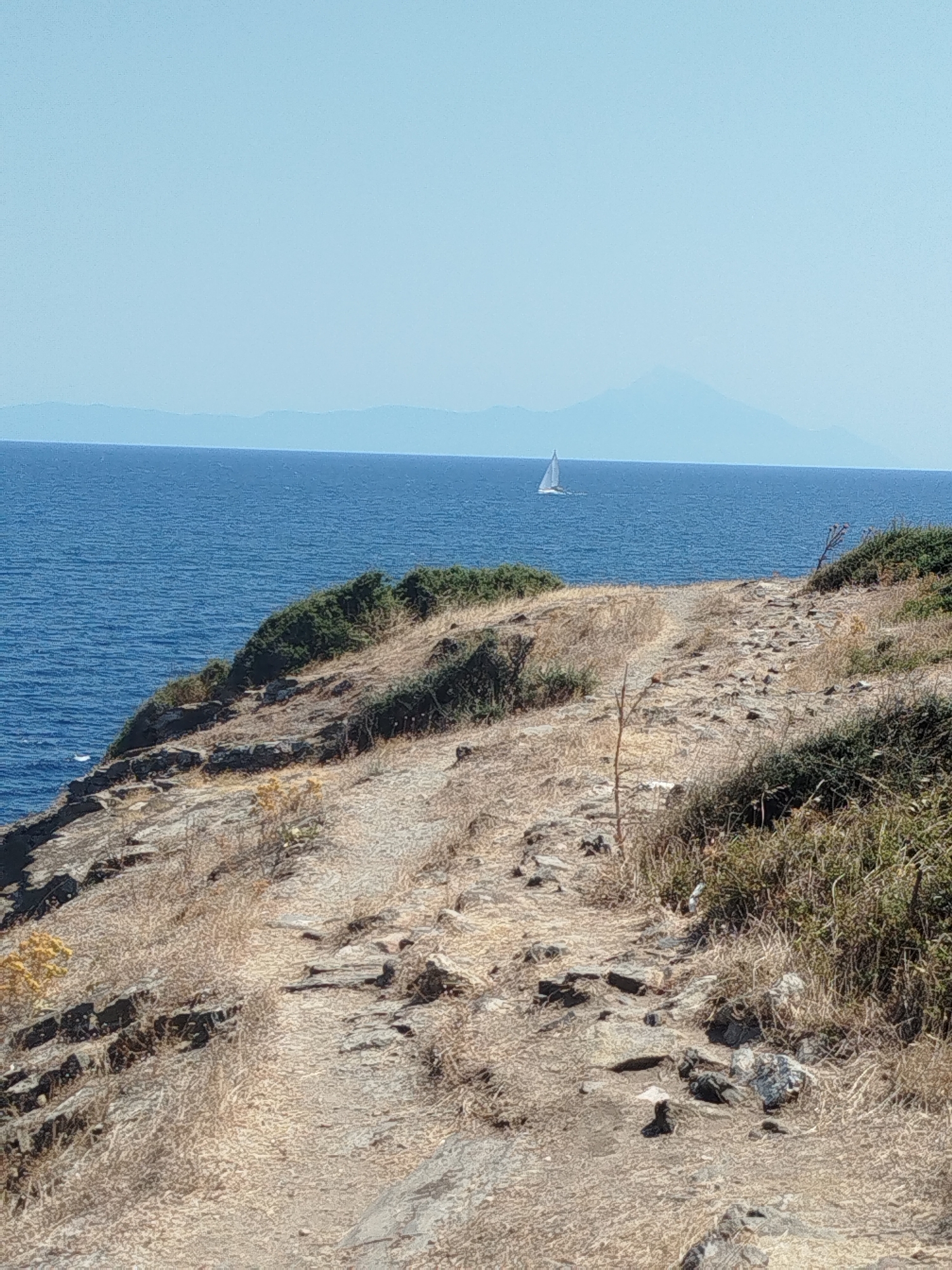 A sailboat is visible in the distance on a calm blue sea, with a mountainous horizon under a clear, pale blue sky. In the foreground, a rocky, dry, and overgrown path winds up a hillside overlooking the sea. The hillside is covered in sparse, dry vegetation and scattered rocks. The overall scene depicts a coastal landscape in Sithonia Municipal Unit, Greece.