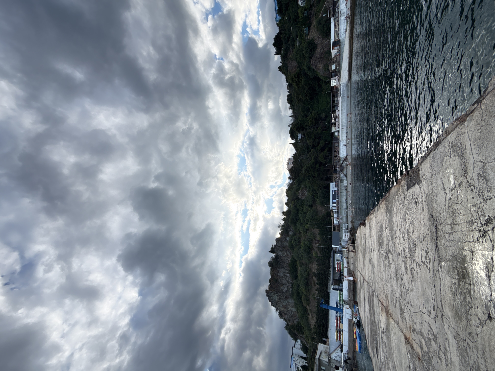 This image depicts a coastal scene in Partenit, Ukraine, likely near the port area. A concrete pier extends into dark, rippling water. The pier's surface is weathered and cracked. On the left side of the pier, a few small boats are moored. Beyond the pier, a coastline is visible, characterized by a rocky hill covered in dense green vegetation. Several buildings, some white and modern, others more utilitarian, are situated along the base of the hill, adjacent to a concrete embankment. A beach area is apparent with scattered individuals. The sky above is dramatic, filled with a mixture of heavy gray clouds and patches of bright blue sky, suggesting changing weather conditions. The overall impression is one of a working port with recreational elements, set against a backdrop of natural coastal topography.