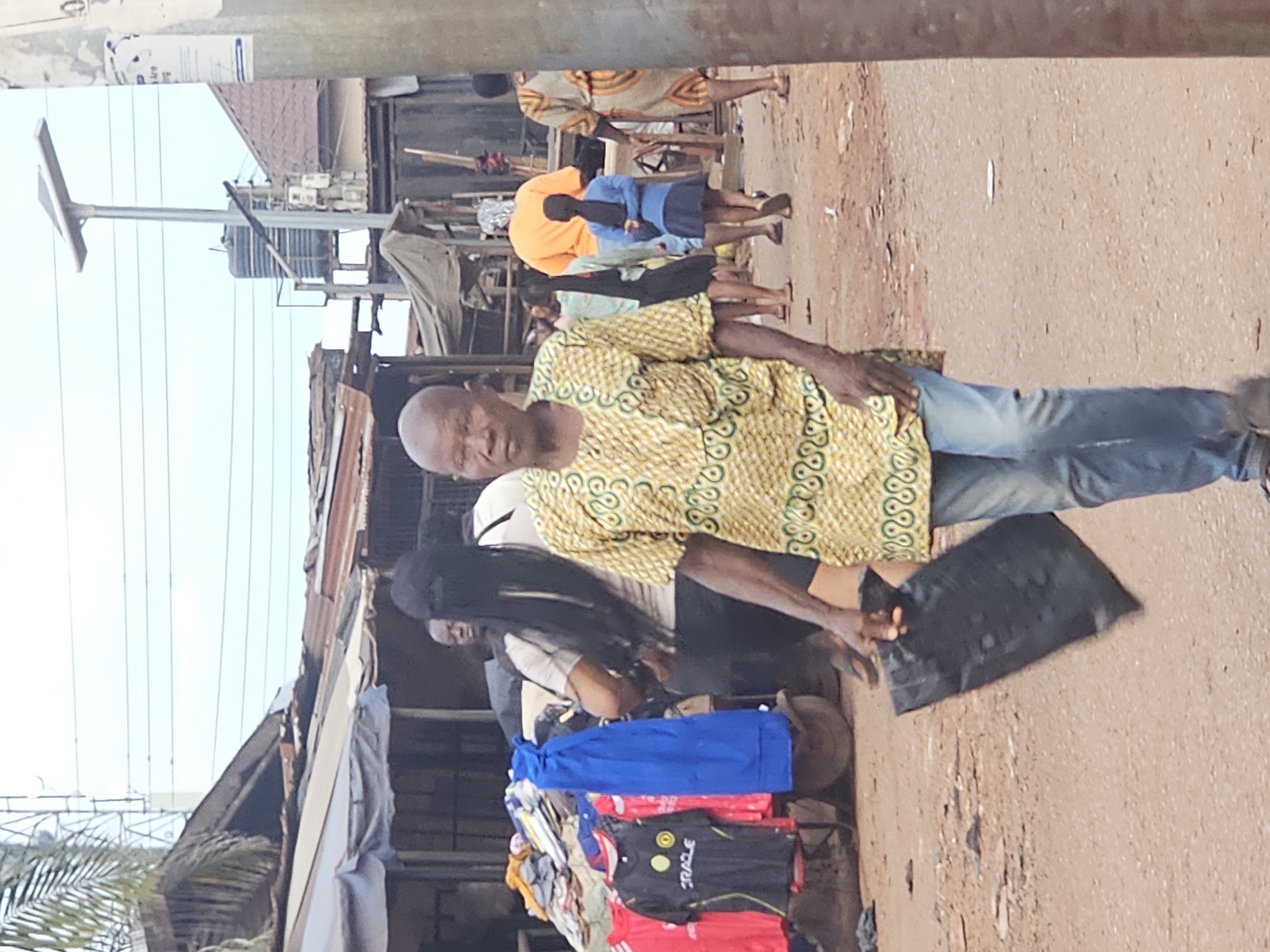 In an outdoor market scene in Unknown, Unknown, a bald man with a light brown complexion walks towards the camera, holding a black plastic bag in his right hand. He is wearing a yellow, green, and white patterned dashiki and light blue jeans. Behind him, a woman with long black hair wears a grey t-shirt and dark pants. Further in the background, a person in an orange t-shirt and a child in a blue shirt and skirt are visible. To the left, a cart displays various colorful t-shirts, including red and black ones. The environment appears to be a dusty street lined with market stalls and overhead wires, with a solar-powered street lamp and a water tank visible in the distance.