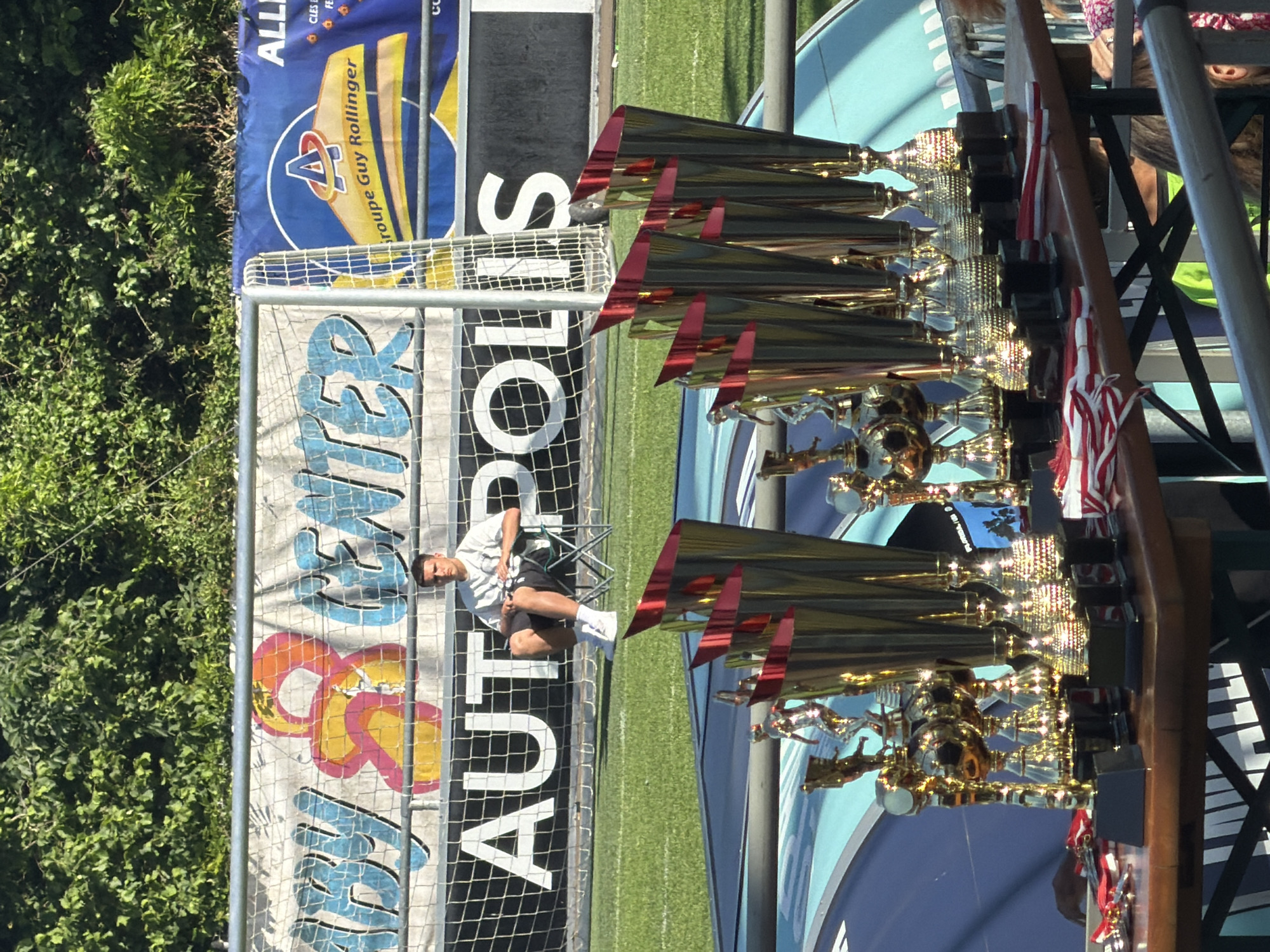 Here is a forensic description of the image:
The image depicts an outdoor scene, with a soccer field, trees, banners, and trophies. In the background, there's a green field and trees on the left side. A banner is visible, with text "ALL" and "Groupe Guy Rollinger" and an "A" logo. Adjacent is a net with the words "AUT POLIS" vertically printed. A man sits on a chair behind the net. In the foreground, there are multiple trophies arranged on a table. Red and white striped ribbons decorate some of the trophies. The location appears to be in Fentange, Luxembourg, based on the banner information and OCR text.