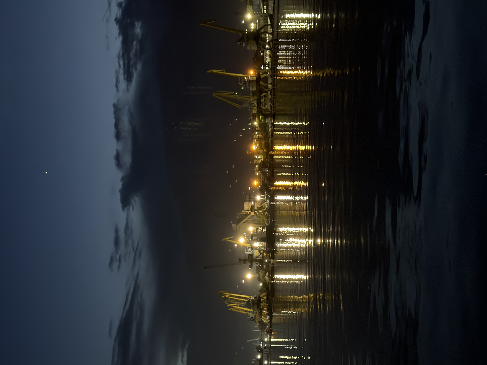 This nighttime image captures a bustling port scene in Burgas, Bulgaria. The primary subjects are numerous large industrial cranes, visible as dark silhouettes and highlighted by bright artificial lights. These cranes, some appearing yellow and green, are positioned along a waterfront.
The lights from the port structures and cranes cast brilliant, elongated reflections across the calm surface of the water, creating shimmering vertical streaks of yellow and white illumination. The sky above is a deep blue-grey, partially covered by dark, textured clouds. A single faint point of light, possibly a star or distant object, is visible in the upper left sky.
The scene suggests active industrial operations, likely involving shipping and cargo handling, continuing through the night. The calm water implies fair weather conditions despite the cloud cover. The strong artificial lighting indicates a functional, working port environment. No specific human activity or text is visible in the frame.