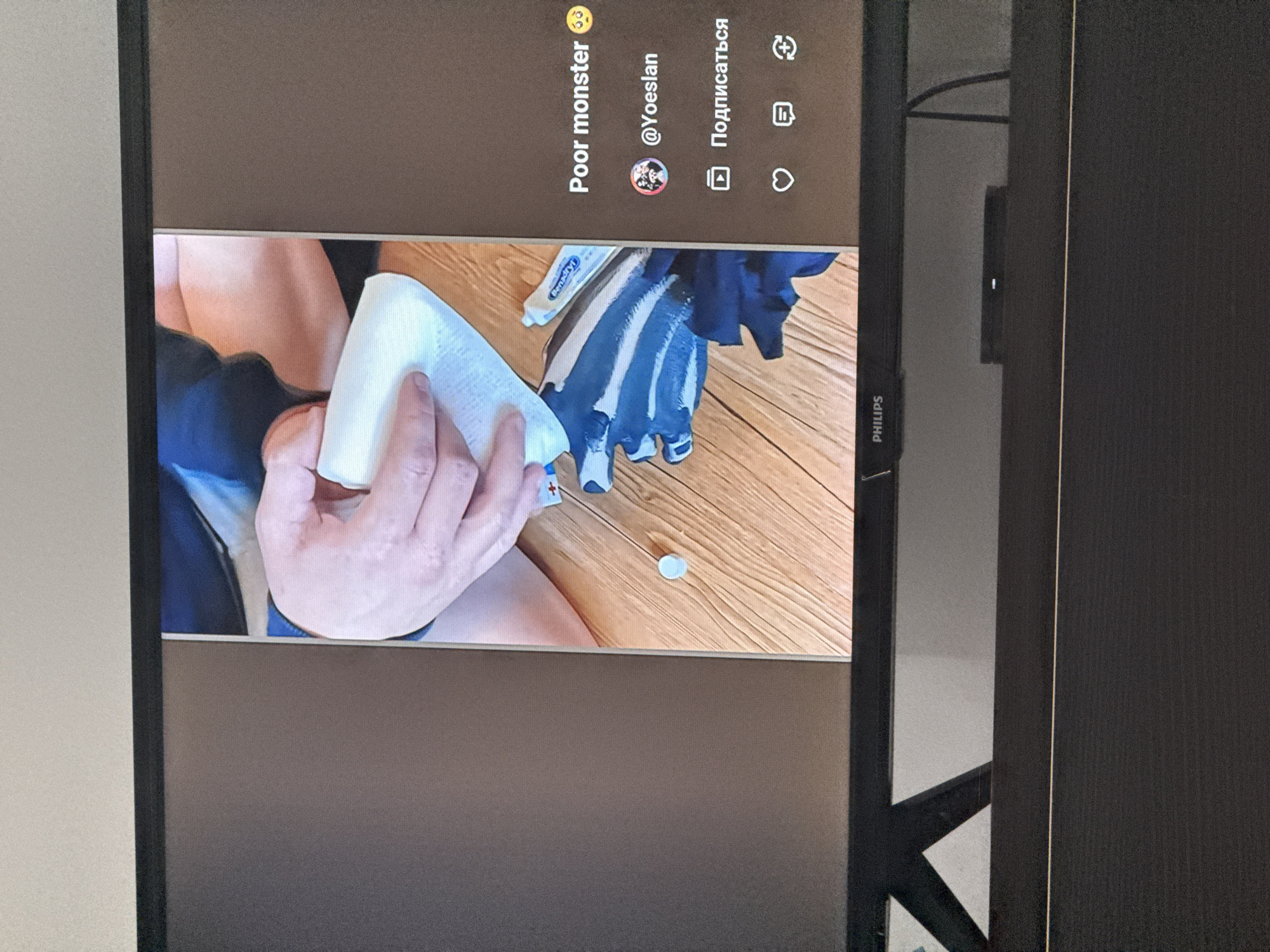A screen displays a video with a user interface overlay. The video shows a person's hands holding a roll of white paper, possibly toilet paper. A tube of "Benadryl" is visible next to the hands. Below the video, the text "Poor monster 🥺" is displayed, followed by "@Yoeslan". Below that, there is a button with a play icon and the Russian word "Подписаться" (Subscribe). Further down, there are icons for like, comment, and share. The screen is positioned on a dark furniture surface. The environment appears to be indoors. There is no indication of the city of Chișinău, Moldova.