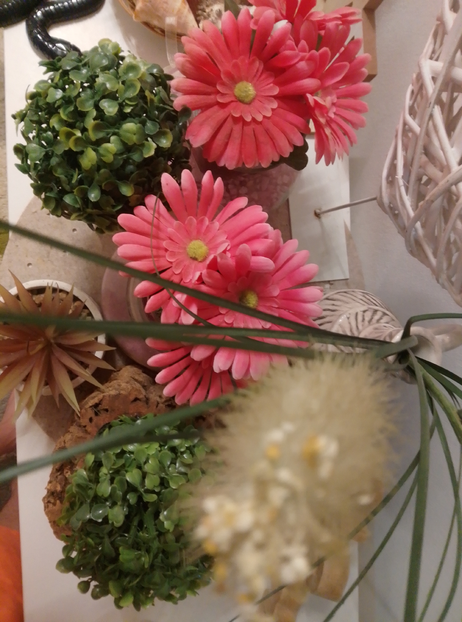 The image shows a top-down view of several decorative items, likely arranged on a shelf or table. On the left side, a dark, segmented object, possibly a snake or a similar decorative piece, is visible. To its right is a round, artificial green plant. In the center, several bright pink artificial gerbera daisies are arranged in a cluster, with their yellow centers clearly visible. Below these flowers, a small white pot contains a spiky, light brown artificial plant. Further down, another small artificial green plant sits on a piece of textured brown material, possibly cork. In the background, a white woven decorative item is partially visible on the right. The overall impression is of a still life arrangement of artificial plants and flowers. No specific location in Wedel, Germany can be determined from the image.