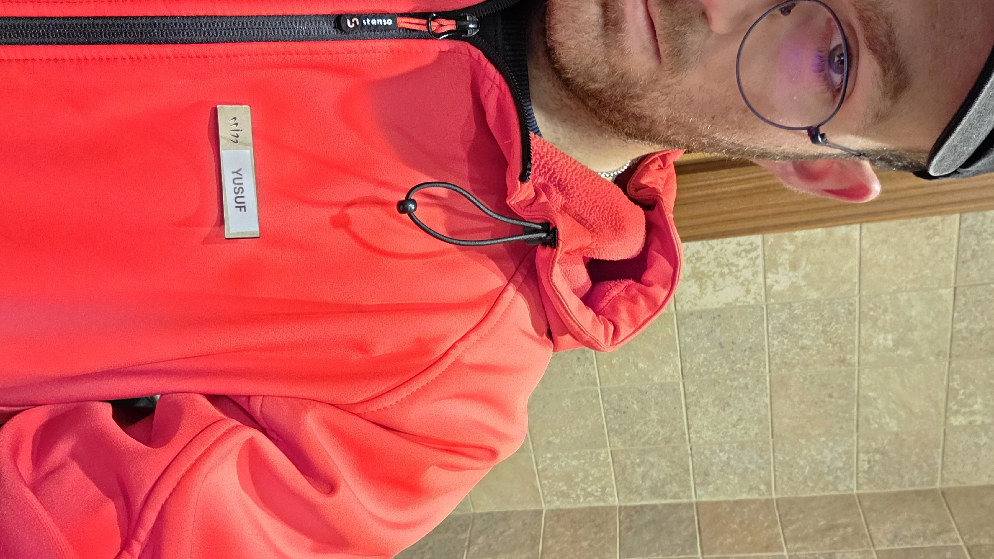 A close-up, slightly rotated image shows a person from the shoulders up, wearing a vibrant red jacket. The jacket features a black zipper down the front, with a zipper pull displaying a white "stemso" logo. A light-colored rectangular tag is sewn vertically onto the jacket's chest, bearing the name "YUSUF" in black capital letters. Above the name, a small stylized symbol is also present on the tag.
The jacket has an adjustable neckline or hood with a black elastic drawstring and a plastic stopper. The person has a light beard and is wearing round, thin-rimmed glasses, with one eye clearly visible reflecting a subtle purple hue. A portion of a black cap is visible on their head.
The background shows light-colored square floor tiles with a textured surface, transitioning to a horizontal wooden trim along what appears to be a wall or baseboard. The lighting is bright, suggesting it is daytime and indoors. The individual is posing for the camera, with no discernible specific activity taking place.