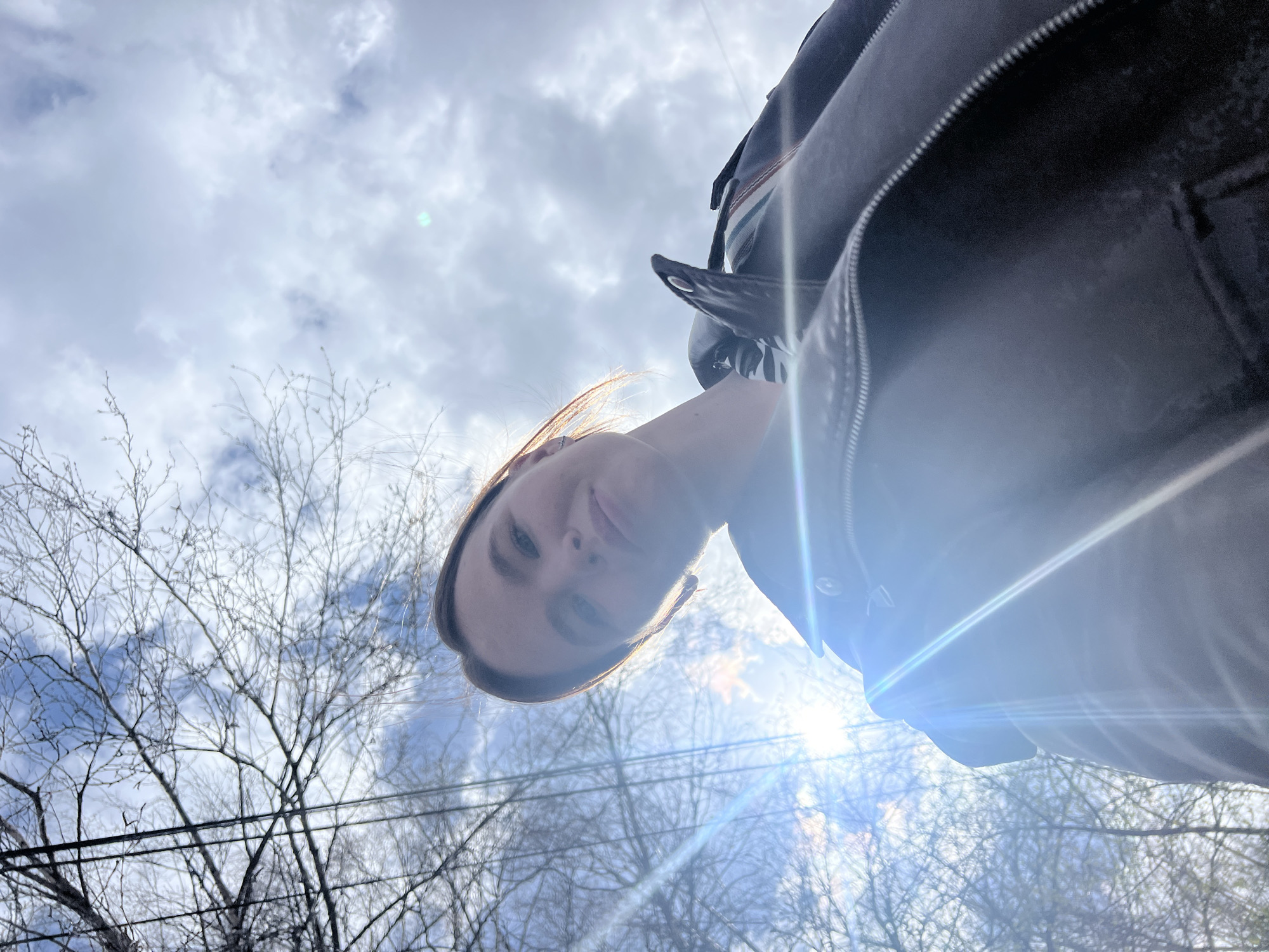 An upward-facing outdoor shot from Chaikovsky, Russia, features a young woman looking down into the camera. Her face, with a neutral to slight smile and light brown hair, is positioned in the lower-center, angled towards the right. She wears a dark jacket or hoodie, visible along the right edge and on her arm, where a black and white striped hair tie and a ring are also seen.
The background is a bright, partially cloudy sky with patches of blue. The sun, located in the lower right, creates significant lens flare and bright light streaks across the image, indicating a sunny daytime. Bare tree branches, suggesting late winter or early spring, are silhouetted against the sky on the left and upper sections. Several dark utility lines or power cables traverse the mid-left portion of the frame horizontally. The overall mood is casual and bright, captured from a low angle. No text is visible.