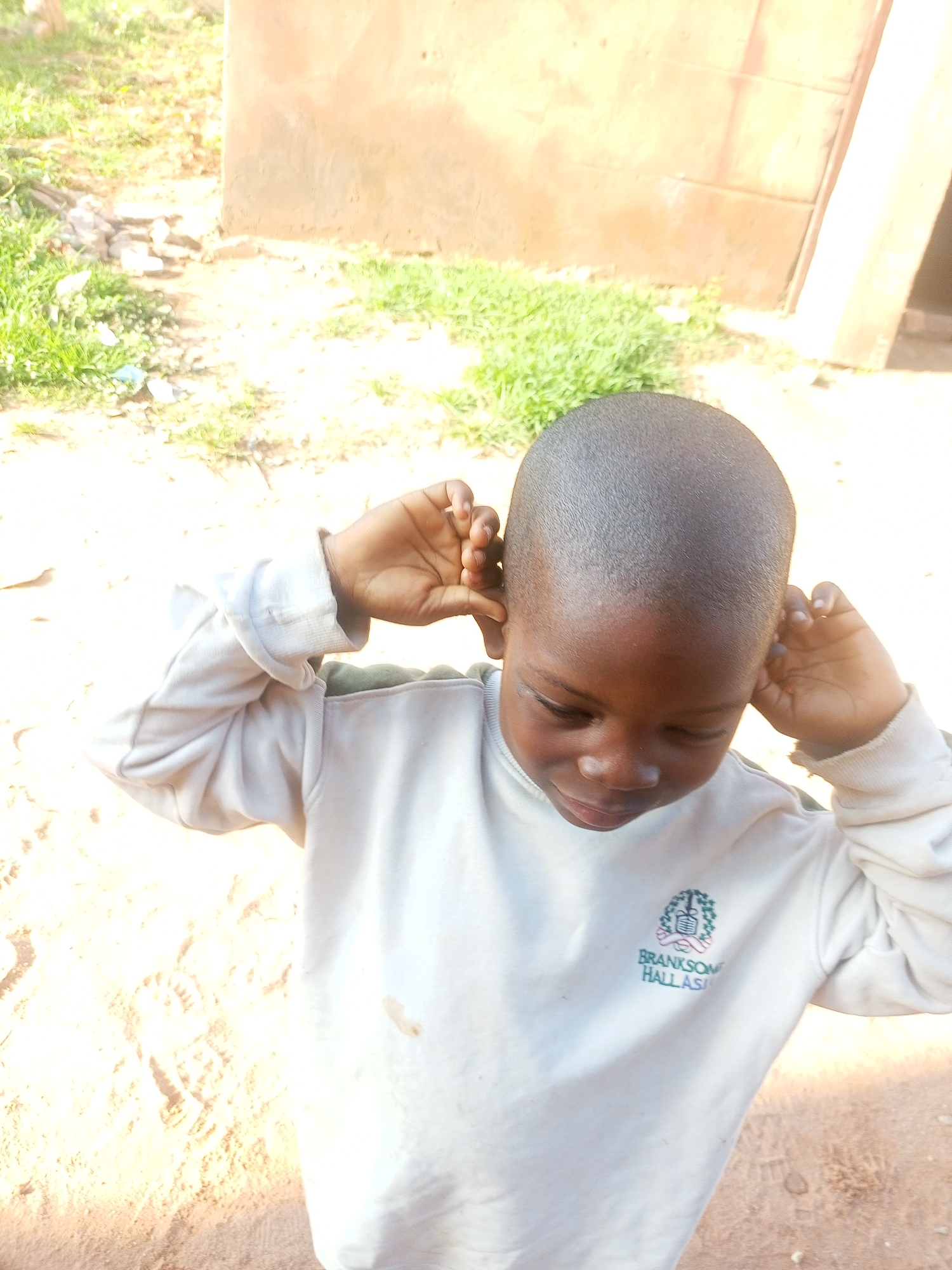 A child with dark skin and a shaved head, looking down with closed eyes and a slight smile, is shown from the chest up. They are wearing a light-colored long-sleeved sweatshirt with a green and blue emblem on the left breast that reads "BRANKSON HALL ASIA." The child's hands are raised, with fingers touching their ears. The background consists of sandy ground with some footprints, sparse green grass, and a light brown wall with a darker rectangular section, possibly a door or window frame. The lighting suggests it is daytime, with bright sunlight casting shadows. This scene is located in Tempure, Nigeria.