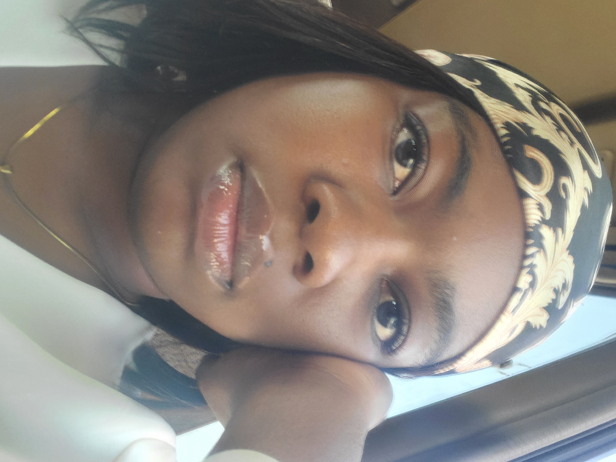 A medium close-up shows a young woman with dark skin and dark hair, wearing a patterned headscarf. Her head is tilted, and her face is toward the camera, with her right hand resting under her chin. She has dark eyes, dark eyebrows, and glossy lips. A thin gold necklace is visible on her neck. The background is blurry and suggests an indoor setting. The location, Gwagwada, Nigeria, cannot be confirmed from the image alone.