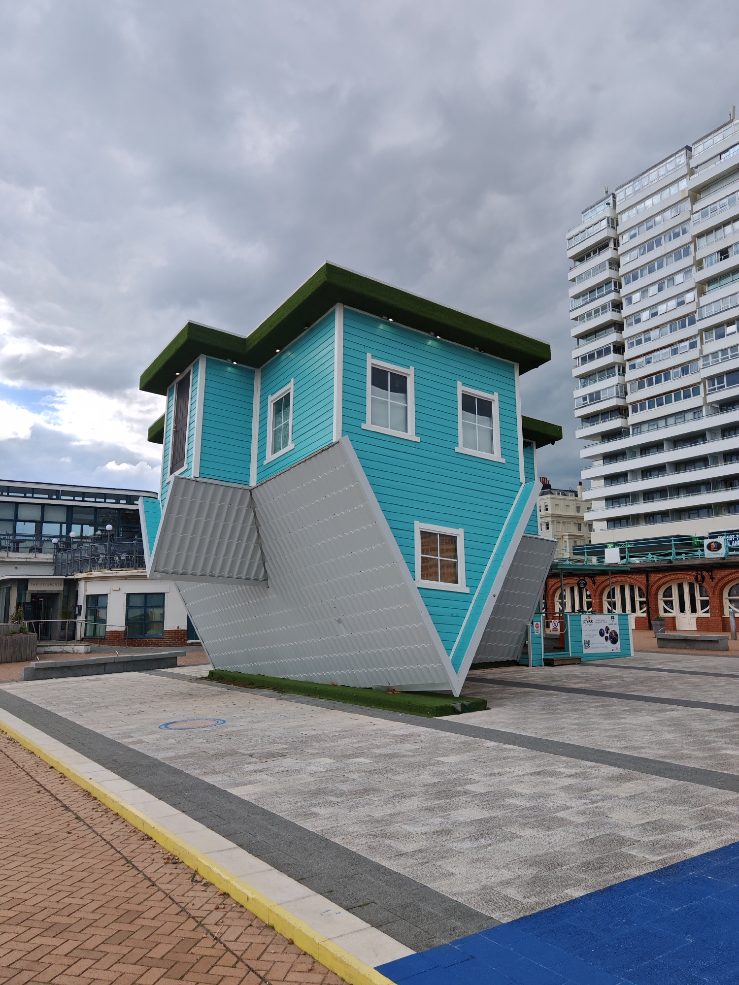 The image shows an upside-down house, painted blue with white trim, in an outdoor plaza. The "roof" rests on the ground, covered with gray shingles, and the "foundation" points skyward with artificial grass. The house is surrounded by paving stones and walkways with gray and yellow curbs. A tall, white building with many windows rises behind the house, and smaller brick buildings are visible in the background. The sky is overcast with gray clouds. The image suggests the location is likely the Upside Down House in the city of Reigate and Banstead, United Kingdom.