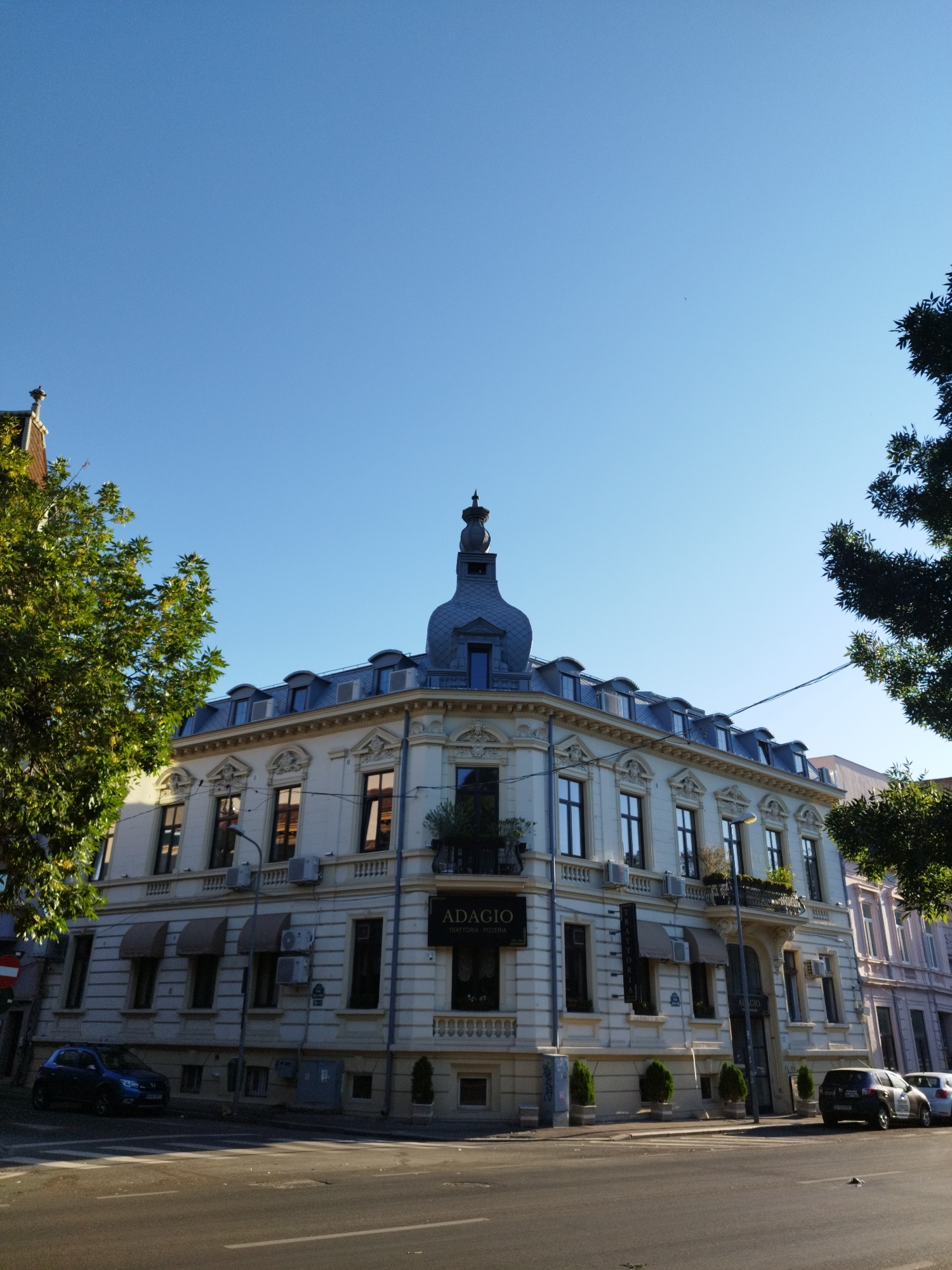 The image shows a corner building in Bucharest, Romania, under a clear blue sky. The building, with its ornate facade and mansard roof, appears to be an older architectural style. A sign on the building reads "ADAGIO TRATTORIA - PIZZERIA". Trees frame the scene on the left and right, casting shadows on the building and the street. Several cars are parked along the street, and a pedestrian crossing is visible in the foreground. The overall impression is of a sunny day in a European city.