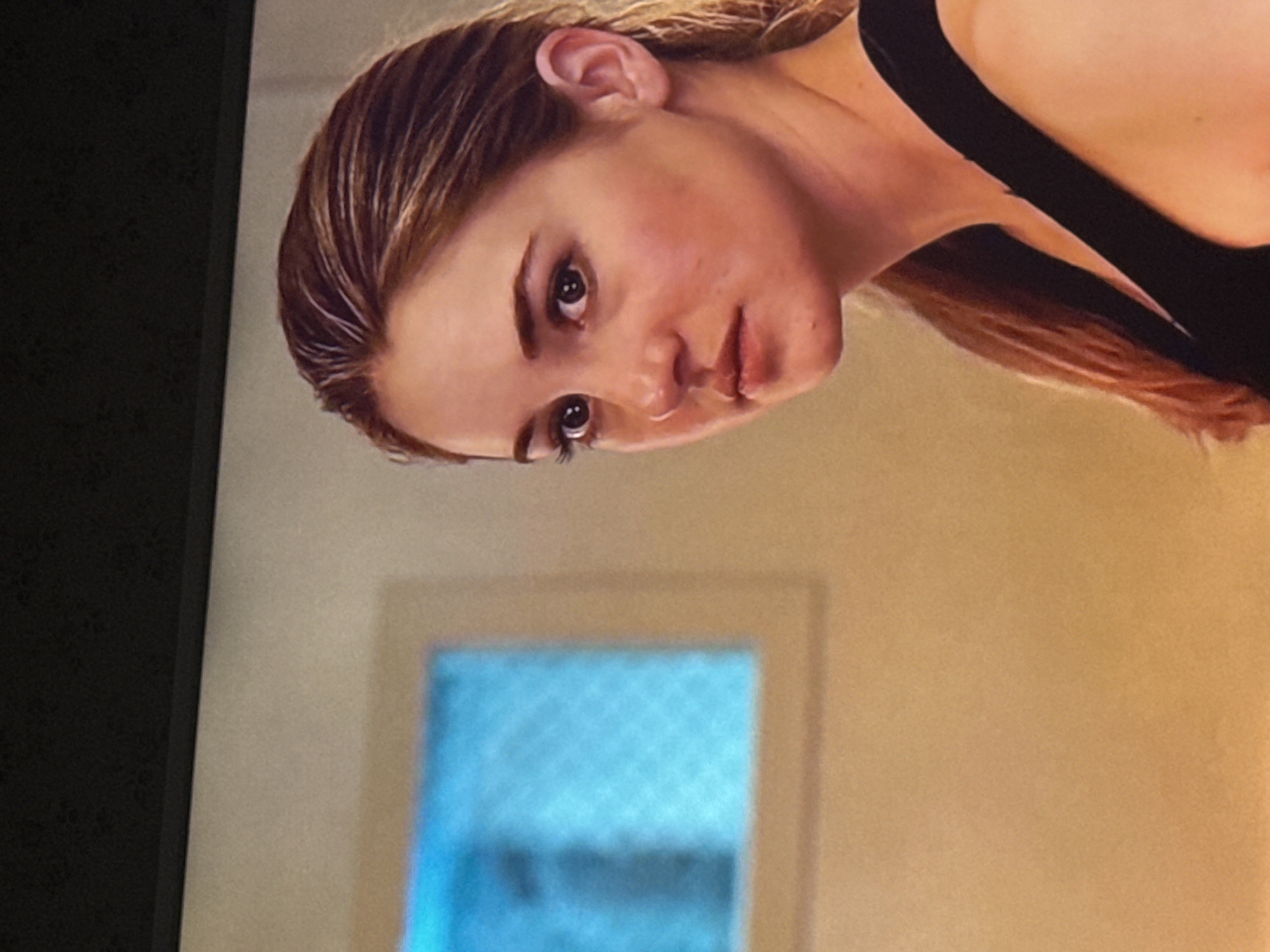 A close-up shot of a woman with light brown hair, wearing a black tank top. The image is slightly rotated counterclockwise. Behind her, a wall with a rectangular window or door is visible. The location of the scene in the city of Mytishchi, Russia cannot be determined from the image alone.