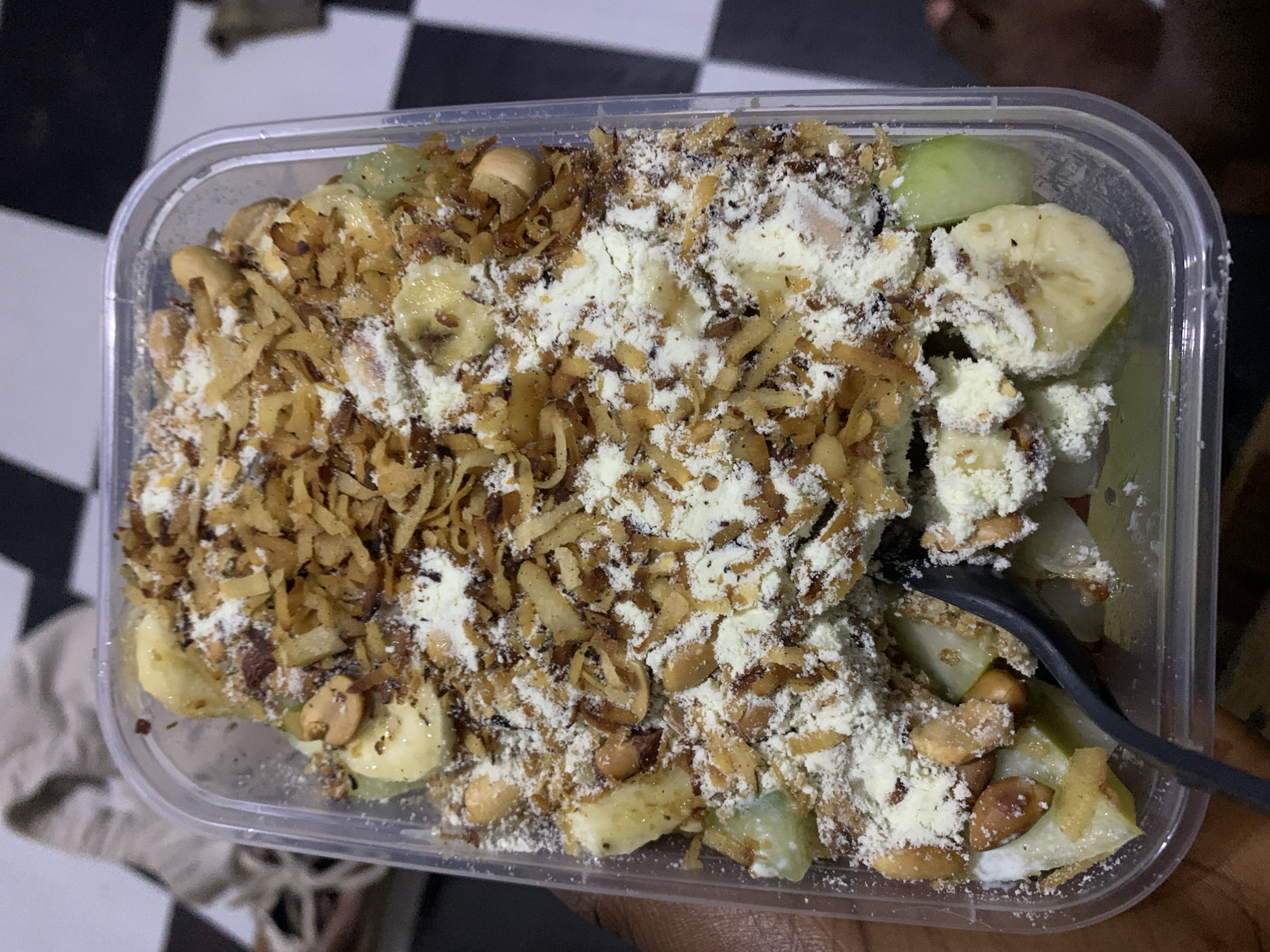 The image displays a close-up, overhead view of a clear plastic rectangular container filled with a dessert or snack. The container holds a colorful mixture of ingredients, dominated by various toppings.
Prominently featured are sliced bananas, identifiable by their light yellow color and characteristic shape, scattered throughout the mixture. Interspersed with the bananas are pieces of green fruit or vegetable, possibly green apple or cucumber, adding a fresh contrast. A significant portion of the surface is covered with a generous amount of finely shredded, golden-brown, crispy topping, which appears to be fried coconut flakes or a similar fried garnish, providing a crunchy texture. Peanuts are also visible, adding another layer of texture and nutty flavor.
A substantial amount of white powdered substance, likely milk powder or a sweet dusting, is sprinkled over the entire dish, creating a snow-like effect and contributing to the overall appearance. A dark-colored spoon is partially visible on the right side of the container, suggesting the dish is ready to be eaten or is in the process of being consumed. A hand is partially visible at the bottom right, holding the container.
The background features a distinct black and white checkered pattern, likely a floor or tabletop, indicating an indoor setting. The lighting suggests ambient or artificial light. No visible text is present in the image. The scene captures a prepared dessert or snack, ready for consumption in Rafin Par, Nigeria.