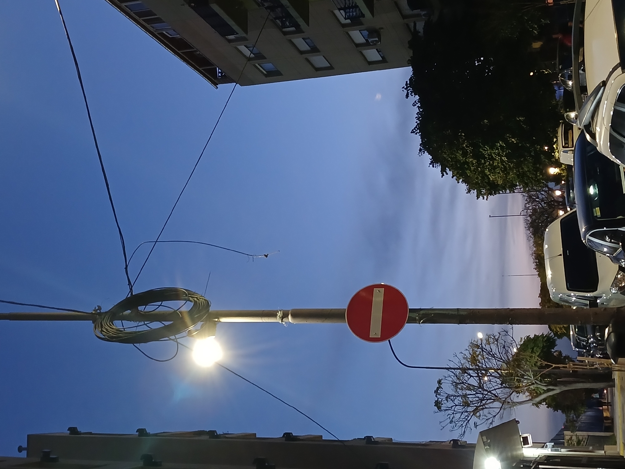A low-angle shot looks up at a street in Buenos Aires, Argentina at dusk. A building looms at the top left, with a dark green tree and parked cars visible on the right. The sky is a clear blue near the building, transitioning to a cloudy, dusky hue. In the foreground, a street lamp post holds a coil of wires, a lit bulb, and a red and white "Do Not Enter" sign. The base of the lamp post is partially obscured by the top of a building.