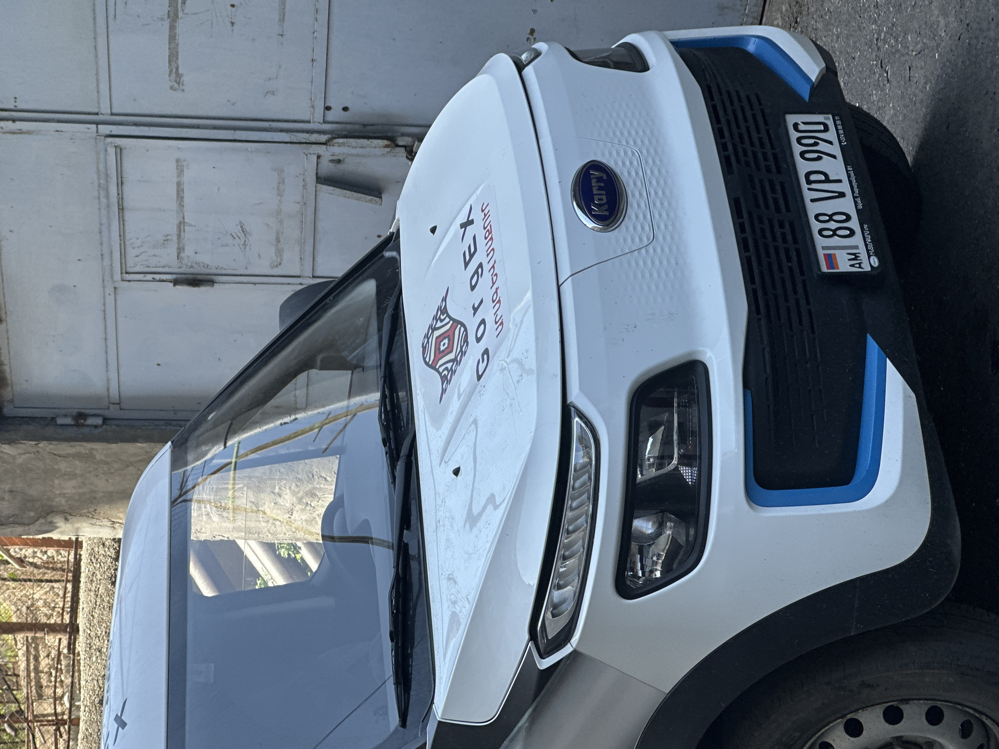 The image shows the front of a white Karry electric vehicle, parked near a metal rolling door, likely a garage. The vehicle features blue trim accents on its front end and a "GorgEX" logo, advertising fast and clean services, plastered on the hood. The license plate reads "88 VP 990" with the "AM" country code, indicating Armenia. Text on the license plate also indicates it is located near Babajanyan 81 in Avan, Yerevan.