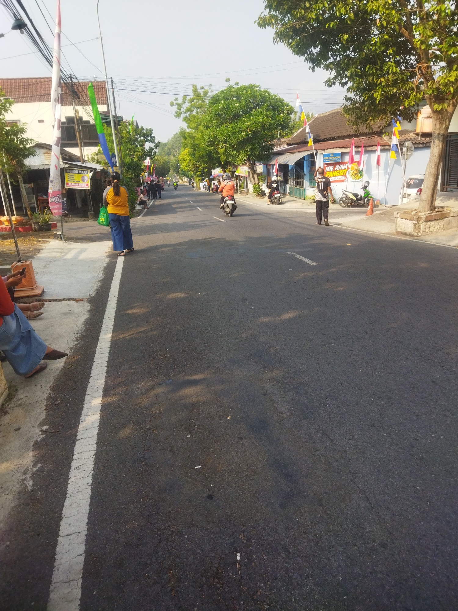 In a street scene in Wonogiri, Indonesia, a road runs through a built-up area with shops and houses lining both sides. The road surface is asphalt, marked with a white line indicating a lane divider. Along the side of the road, a sidewalk is visible, with a few individuals sitting on it. One person, wearing a blue skirt and an orange top, is holding a green bag. Another person, with legs crossed and wearing a blue skirt, is seated, appearing to use a mobile phone.
On the road, several motorbikes are present. One motorbike is ridden by a person wearing an orange jacket, while another is parked. A person in a black shirt with white numbering is walking on the road. People are also seen walking in the distance. Decorative flags in red, white, blue, and yellow are displayed along the street, attached to poles and the buildings. Trees with green foliage are scattered throughout the scene, with one prominent tree on the right side of the image. The sky is clear and bright, suggesting it is daytime. The general atmosphere appears to be that of a busy street in a residential or commercial district.