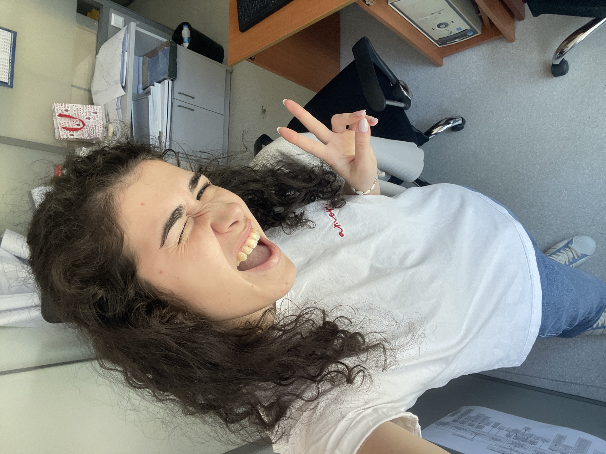 A young woman with dark curly hair is making a peace sign and looking at the camera with a squinted eye and open mouth. She is wearing a white t-shirt with red embroidery on the front, and blue jeans. She has a bracelet on her left wrist. The background appears to be an office setting. There is a desk with a computer on it, a chair, and a filing cabinet. The floor is grey. The exact location in Sofia, Bulgaria cannot be determined from the visual information.
