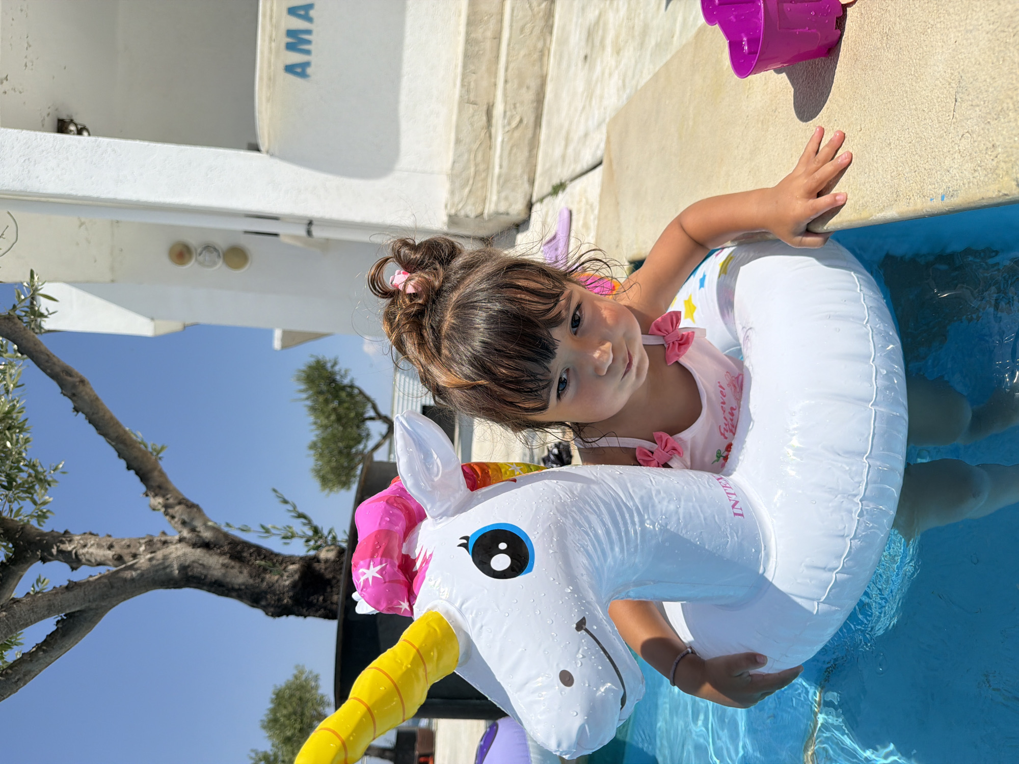 A young girl with dark, curly hair tied up in a messy bun, adorned with pink bows, floats in a bright blue swimming pool. She is wearing a pink swimsuit with a cherry print and the words "Forever fun" visible. She is holding onto a large, white inflatable unicorn that has a rainbow-colored mane and horn. The unicorn has large, black and blue eyes and a friendly smile.
The girl is partially submerged in the water, and the unicorn float encircles her. Her hand rests on the side of the pool, which has a textured, sandy surface. In the background, parts of a white building and stone stairs are visible. A large tree with sparse leaves branches out over the pool. The sky is a clear, bright blue, suggesting it is a sunny day, likely during daytime.
A small, purple plastic bucket is seen near the edge of the pool. The inscription "IN..." is partially visible on the unicorn float. The overall scene depicts a joyful moment of a child enjoying a summer day by the pool in Burgas, Bulgaria.