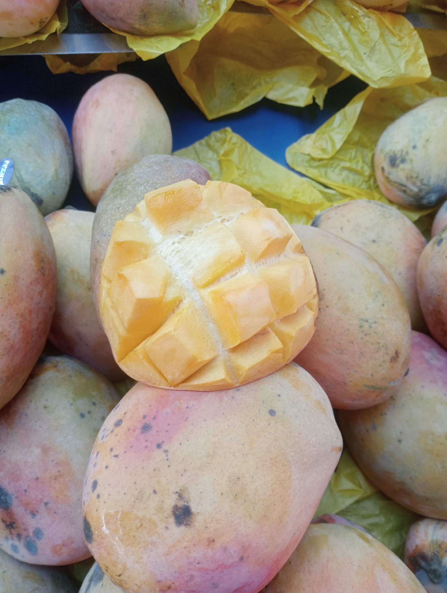 A close-up, high-angle shot shows a pile of ripe mangoes, some whole and one cut in half with its flesh cubed. The mangoes are a mix of yellow, orange, and red hues, with some displaying small dark spots on their skin. The cut mango half reveals a bright yellow-orange interior with a textured surface. The fruits are arranged on what appears to be yellow paper or plastic sheeting, with glimpses of a blue surface in the background, possibly a market stall or display. The lighting suggests an indoor or shaded outdoor setting. The scene is devoid of people or any discernible action. This image could have been taken at the Antalya Central Market (Antalya Hal Pazarı) in Antalya, Turkey, a common location for fresh produce displays.