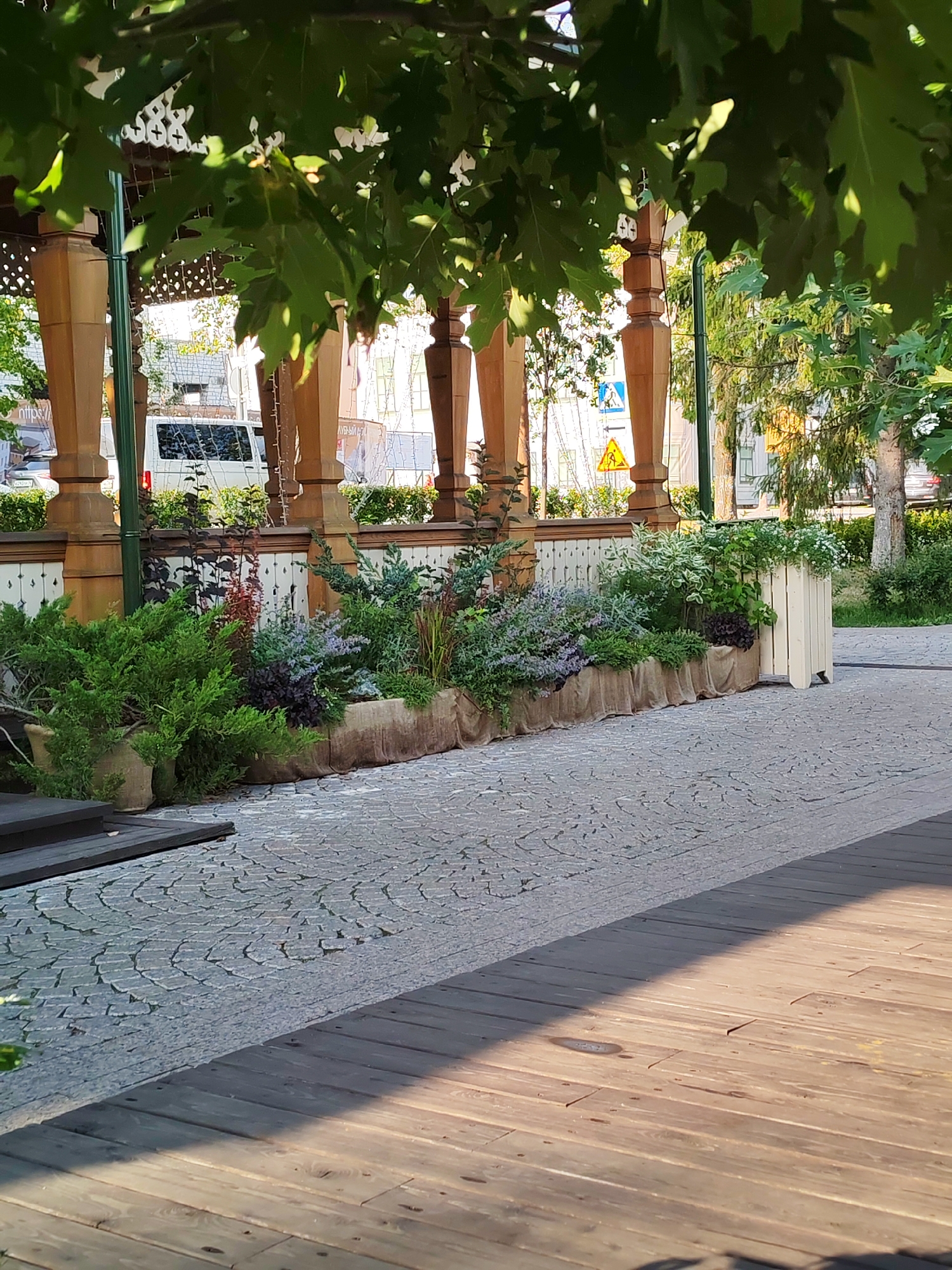 This is a medium shot of an outdoor area with a paved walkway, a wooden deck, and a raised planter box filled with various green plants and some purple flowers. The planter box is covered in burlap. Behind the planter box is a white wooden fence with decorative elements and several wooden columns that appear to be part of a gazebo or covered seating area. In the background, there are buildings, trees, and a white van. The time of day appears to be late afternoon or early evening, as evidenced by the warm sunlight casting long shadows across the wooden deck and the cobblestone pavement. The weather seems pleasant and sunny. There are no people visible in the image. Some text is visible on a white van in the background, but it is too small and blurry to read. There are also yellow and blue road signs visible in the background. The scene is likely a public outdoor space, possibly a park or a cafe patio, in Bol'shie Mkhi, Russia. The overall mood is peaceful and serene, with a focus on the lush greenery and the architectural details of the structure.