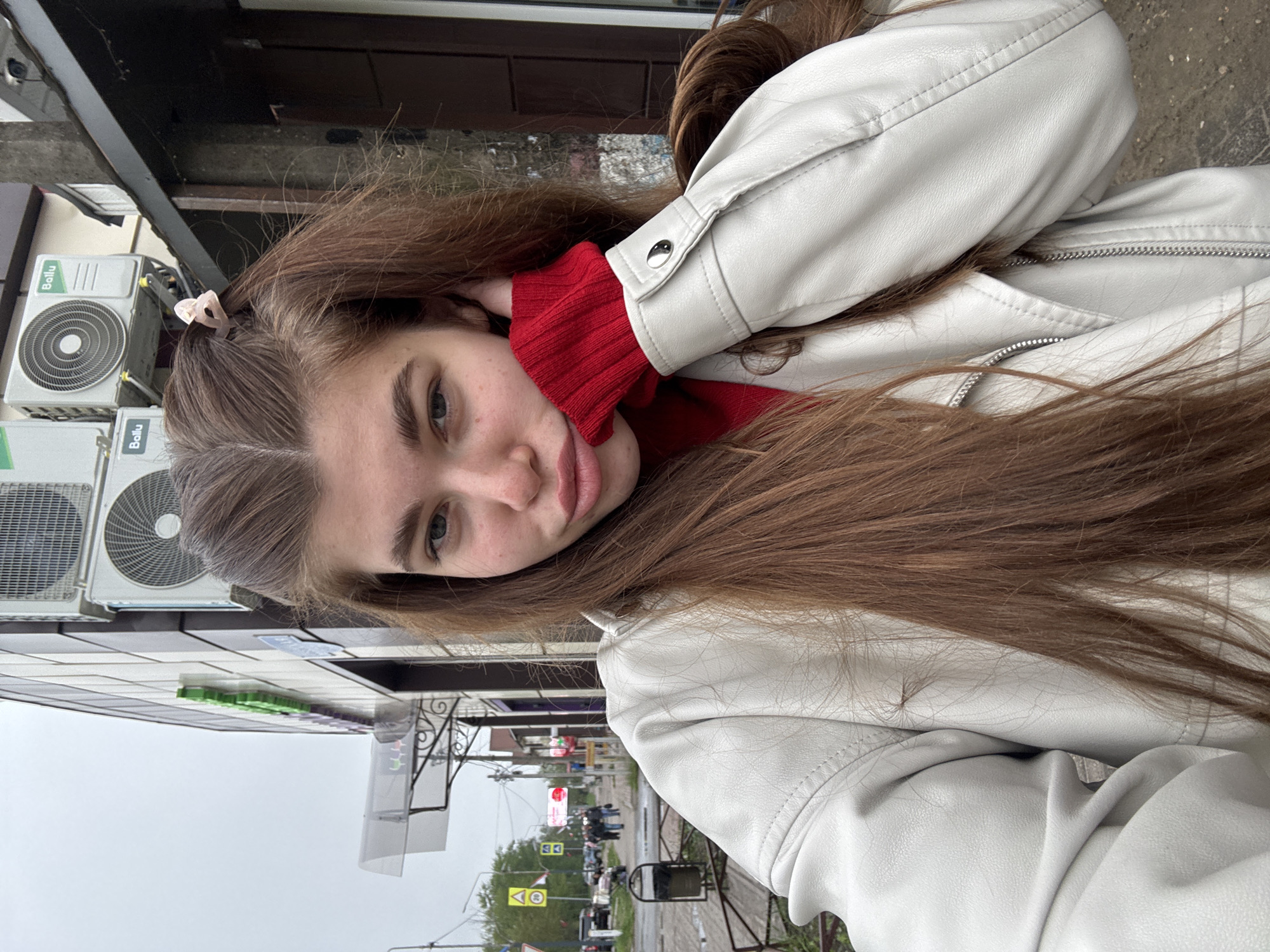 A young woman is photographed in a candid, close-up shot. She is seen in a white jacket over a red garment. Her long, brown hair is visible and a small, light-colored hair clip is in her hair.
The photograph appears to be taken outside, next to a building with visible air conditioning units marked with "Ballu." A street scene with traffic lights and pedestrians is in the blurred background, indicating an urban environment. Based on the presence of Cyrillic text on the signage and the fact that the query mentions Kurovskoye, Russia, it can be inferred that this photo was taken in that city.