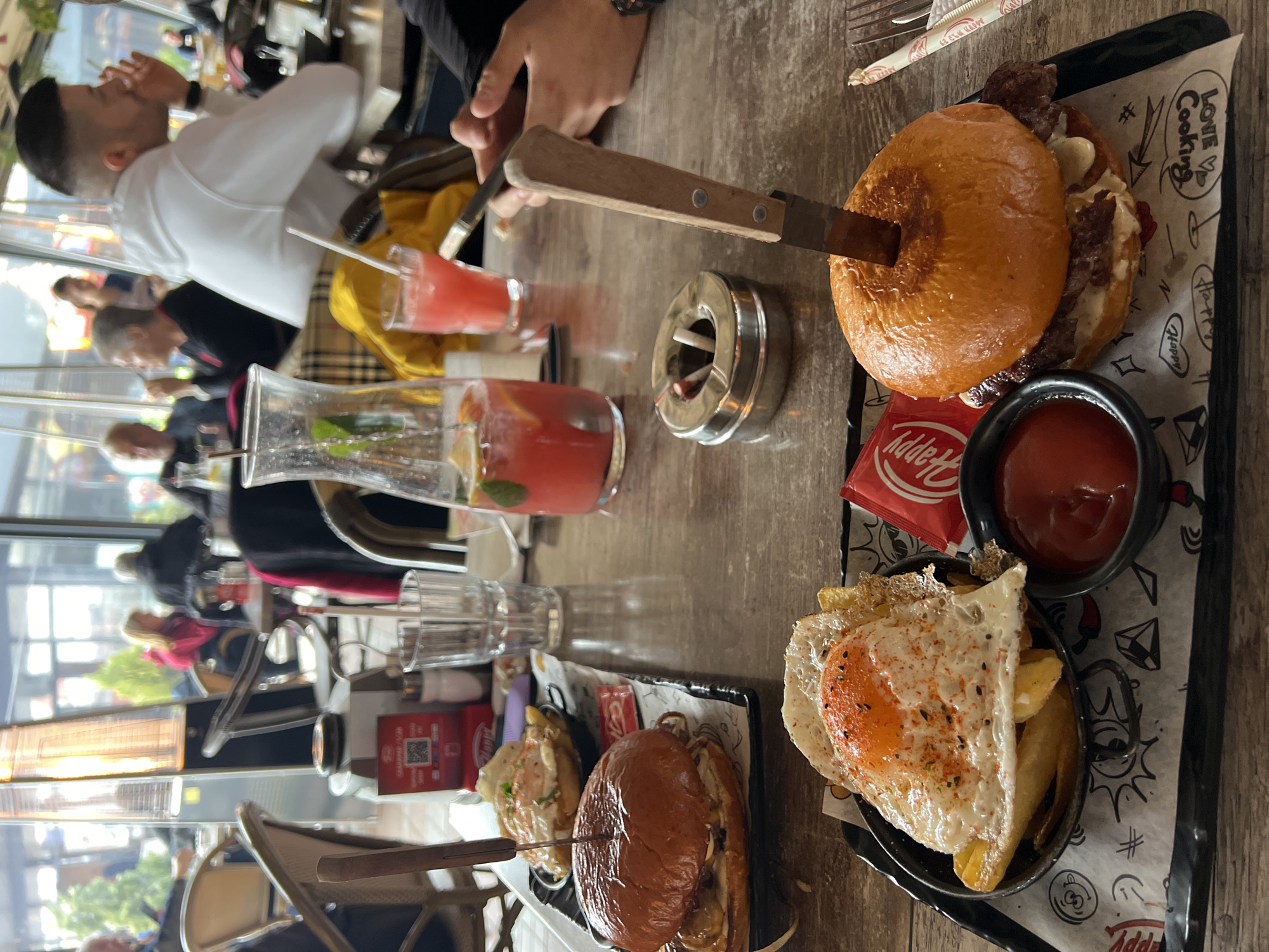 The image captures a dining scene at a Happy Bar & Grill restaurant in Sofia, Bulgaria, characterized by a modern, casual interior with large glass windows suggesting a semi-enclosed terrace. In the foreground, a light-colored wooden table features two black rectangular serving trays. On the bottom right tray, a burger with a glossy brioche bun is held together by a large knife plunged vertically through its center. Alongside it are a small black bowl of ketchup, a red 'Happy' branded packet, and a wrapped fork and straw. The middle-left tray presents another burger, similarly secured by a knife, and a small metal bowl containing French fries topped with a fried egg, seasoned with red flakes and black sesame seeds.
Between the trays, a silver metallic ashtray holds a cigarette butt and a plastic stirrer. Beverages on the table include a clear glass carafe of a red fruit drink garnished with mint and citrus slices, and a tall glass of a similar red drink with a straw. Other table items include an empty clear ribbed drinking glass, a red rectangular stand displaying a QR code, and a small salt shaker.
In the midground, a male individual with short dark hair and a beard, wearing a white hoodie, is seated, looking towards the left, with his right hand near his face. Behind him, a person wearing a yellow jacket is partially visible. The background, out of focus, reveals multiple blurred figures of other patrons seated at tables, visible through the large glass windows. An active outdoor patio heater with a visible flame stands within this area, and green foliage is discernible outside the windows.