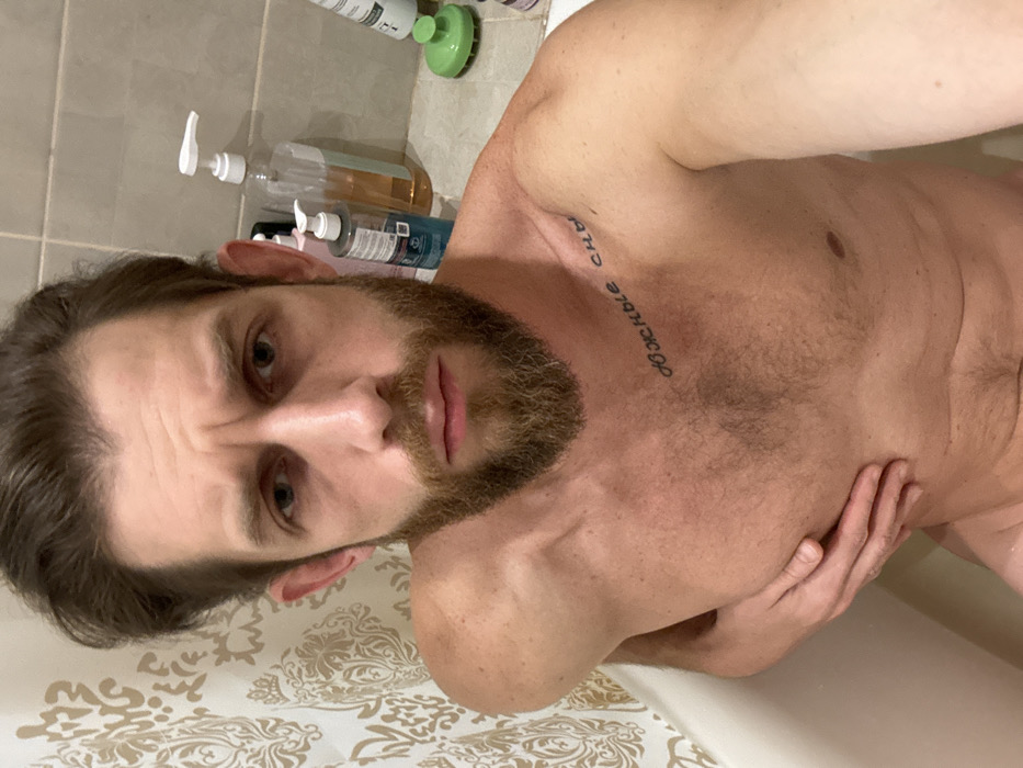 A close-up, angled selfie of a bearded man in a bathroom. He is shirtless, with fair skin and a tattoo visible on his chest. His hair is dark and slightly disheveled. He is looking at the camera with a slight furrow in his brow. In the background, there are various bottles of shampoo and body wash on a shelf. The shower curtain is visible in the foreground.