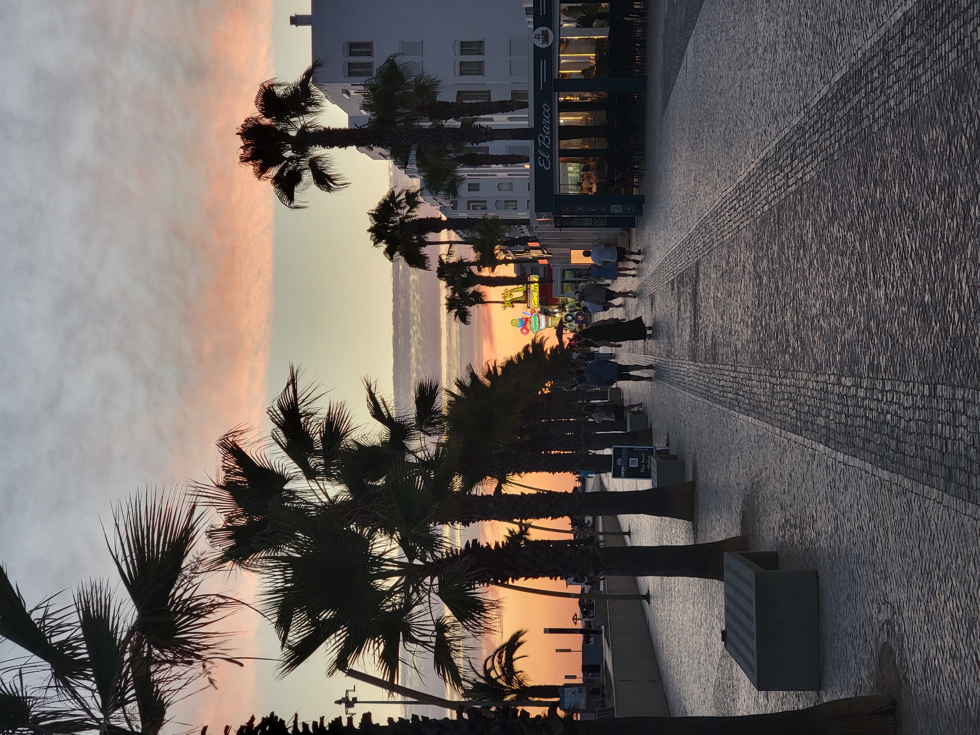 The image shows a sunset scene on a promenade in Quarteira, Portugal, with palm trees lining the walkway. The sky is a mix of orange and pink hues, with scattered clouds. A paved pathway, made of cobblestones, stretches into the distance, with several people walking along it. On the right side of the promenade, a white building with multiple windows stands, and a restaurant with outdoor seating is visible. The restaurant's sign reads "El Barco." There are also several concrete benches along the promenade. The overall atmosphere is tranquil, with the setting sun casting a warm glow on the scene.