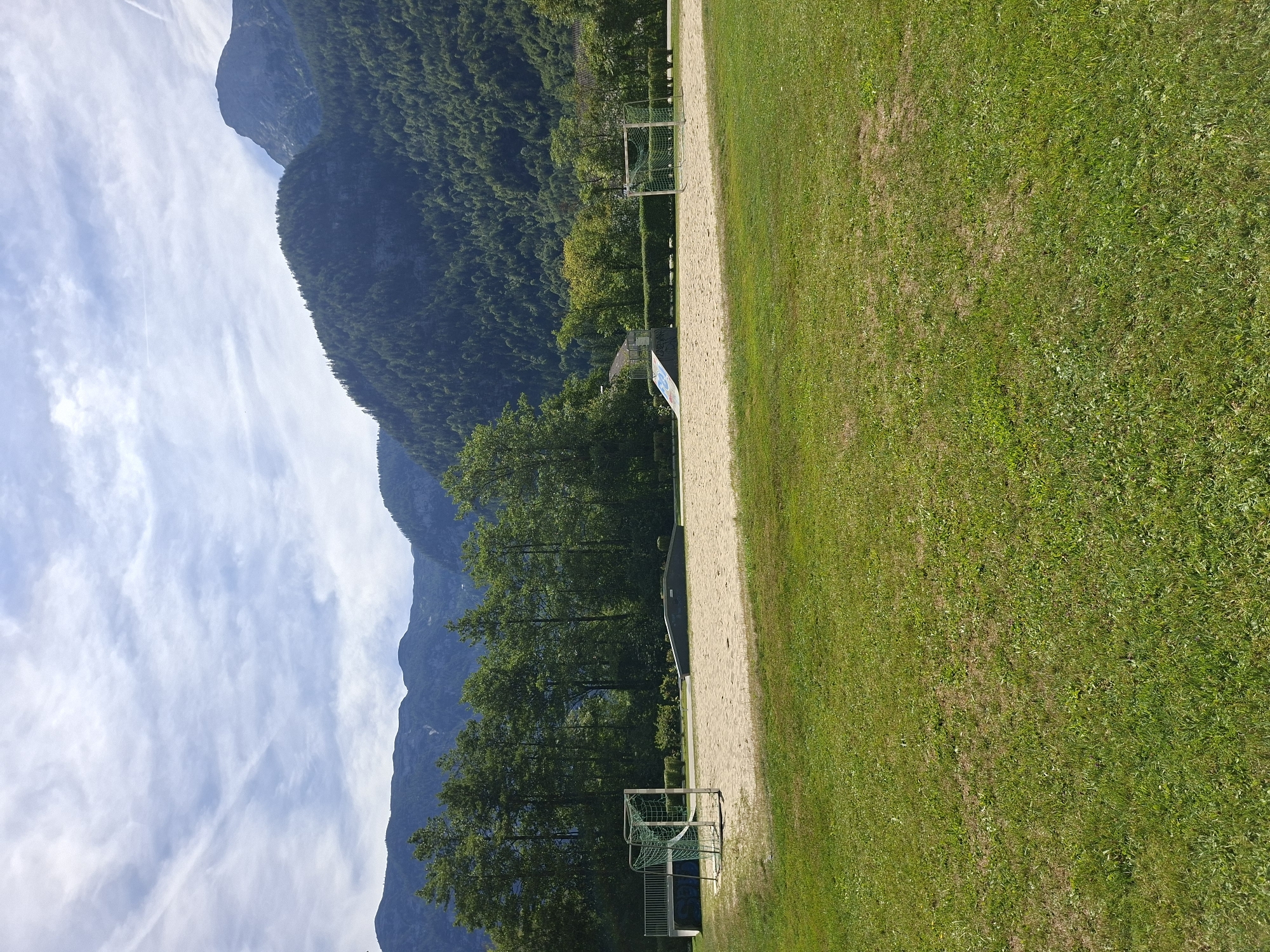 The image depicts a sunny outdoor recreational area located in Obertraun, Austria. In the foreground, a lush green lawn occupies the lower portion of the frame. The midground features a sand court with a small soccer goal on the left and a ramp or skate feature on the left side of the sand. Beyond the sand court, a line of mature trees forms a dense green backdrop. In the distance, imposing mountains rise, their slopes covered in dark green forests, under a bright blue sky with wispy white clouds. The scene conveys a sense of a peaceful, natural environment suitable for outdoor activities.