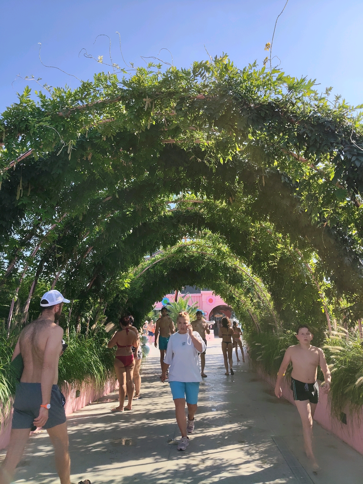 The image shows a pathway under a leafy green archway, possibly in a park or recreational area. Several people are walking along the path. In the foreground, a man wearing a white cap and grey shorts walks away from the camera. Behind him, a woman in a red bikini walks away from the camera. Further down the path, other individuals are visible, some in swimwear. The archway is formed by dense green foliage with some long, trailing vines. Sunlight filters through the leaves, casting shadows on the path. In the background, structures with pink walls and arched entrances are visible. The scene appears to be a bright, sunny day. There is no identifiable information suggesting the location is in Chișinău, Moldova.