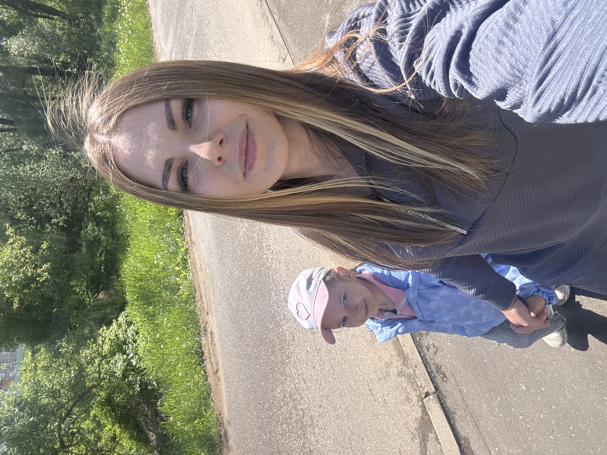 A woman with long, highlighted hair is taking a selfie with a young child. The woman is wearing a blue-gray ribbed top. The child, wearing a pink and blue patterned cap with a heart on it and a blue jacket, is holding the woman's hand. They are standing on a paved path next to a grassy area with trees. The location is potentially Shevlyakovo, Russia.