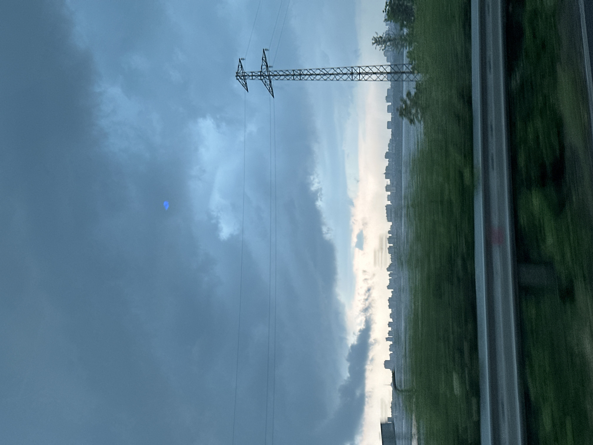 The image, when viewed upright, displays a landscape under a dramatic, overcast sky. Heavy, dark grey and blue-grey clouds fill the upper two-thirds of the frame, suggesting impending or recent stormy weather. In the upper left portion of the sky, there is a distinctive bright blue, irregularly shaped artifact, possibly a lens flare or reflection.
A prominent lattice-structured electrical pylon and its associated power lines stretch horizontally across the mid-ground, silhouetted against the dark sky. Multiple power lines extend from the pylon into the distance. Below these lines, along the horizon, lies the faint, indistinct silhouette of a distant city skyline, characterized by numerous buildings.
The lower third of the image features a multi-lane highway or road, distinguishable by its parallel grey concrete or asphalt strips. Adjacent to the road and partially obscuring the distant city is blurred green foliage, which could be grass or trees. The blurring of the foreground elements suggests the photograph was captured from a moving vehicle.
The scene conveys a sense of transition, likely late afternoon or early evening, under subdued lighting conditions due to the heavy cloud cover. The setting combines industrial infrastructure (pylon, power lines, highway) with natural elements (sky, clouds, greenery) and a distant urban backdrop. There are no people visible, and no discernible text.