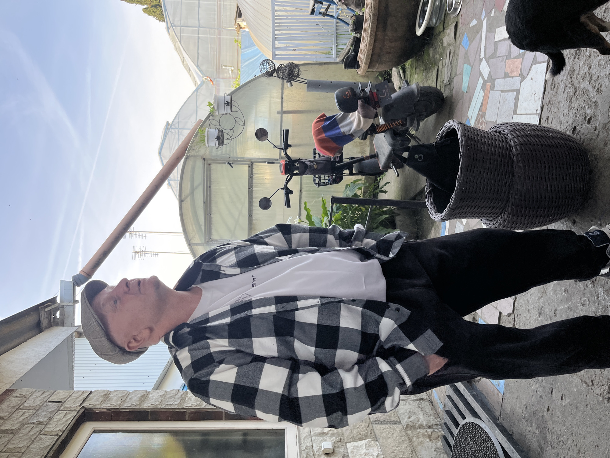An outdoor, rotated image depicts a man standing in what appears to be a garden or courtyard area, with multiple greenhouses visible in the background under a bright, clear sky. The ground is a mix of irregular stone paving and colorful mosaic tiles.
The main subject is a fair-skinned man, seen from the chest up, wearing a black and white plaid flannel shirt over a white t-shirt, black pants, and a grey flat cap. He has short hair and is looking upwards and slightly to his right with a neutral, contemplative expression. His hands are in his pockets. On his white t-shirt, the text "SPRT" is visible.
In the mid-ground, an electric scooter or moped, partially covered with a red, white, and blue blanket or towel, is parked near a wicker basket. Inside the basket, a dark object, possibly a black bag or another animal, is visible. A dark-furred dog is partially seen from behind to the right of the basket, its hindquarters and tail visible. Further back, several potted plants are arranged near the greenhouses. Antennas are visible on a nearby roof. The e-scooter has "LUODAO" partially visible on its side.
The setting suggests a sunny daytime scene, possibly in the morning or late afternoon, given the clear light. The presence of greenhouses, potted plants, and the general rustic paving indicates a rural or semi-rural environment, consistent with a location like Vesyoloye, Russia. No specific activity beyond standing and observing is depicted.