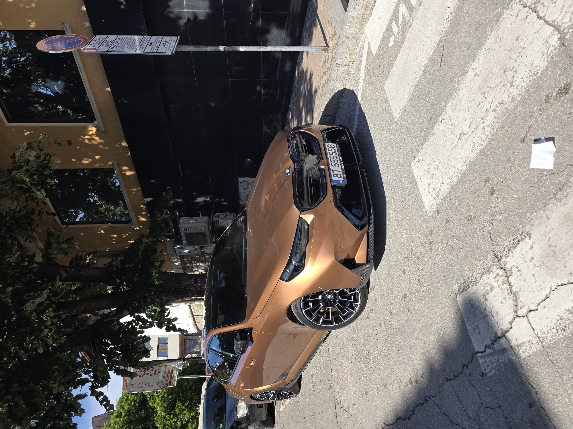 The image depicts a copper-colored BMW parked on a street in Veliko Tarnovo, Bulgaria. The license plate of the car reads "BT 55555B." The car is situated on a pedestrian crosswalk near a building, which has a "No Parking" sign posted. A hotel sign, "Hotel Comfort," is also visible near the car. The background includes trees, a portion of a building, and the street. The lighting suggests it's daytime.