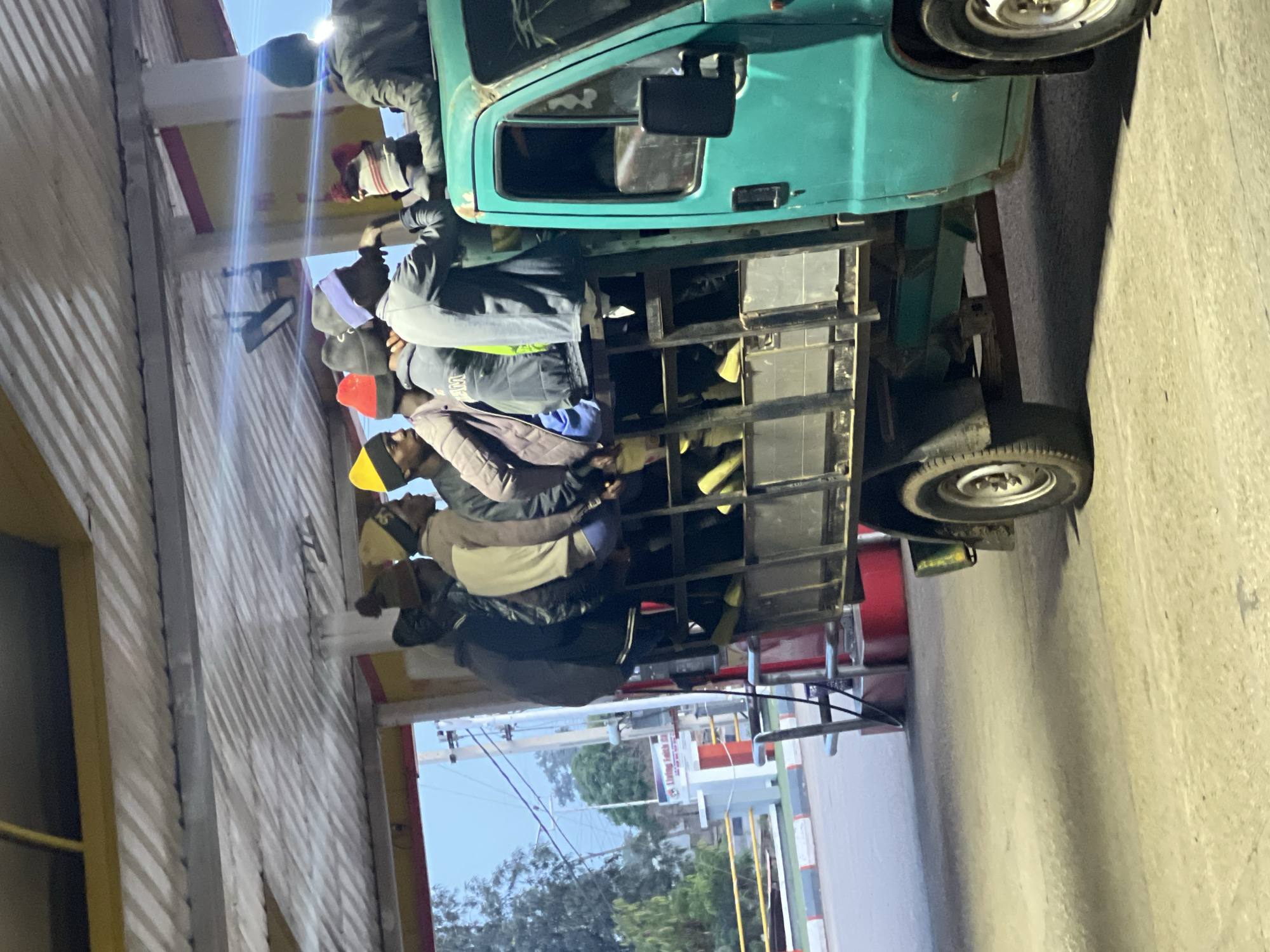 The image depicts an outdoor scene featuring a light-green truck transporting multiple men in its open cargo bed, which is fitted with metal railings or a cage. The truck is positioned on a paved surface next to buildings with corrugated metal siding.
Approximately ten to twelve men are visible in the truck's bed, either standing or seated along the edges. They are dressed in casual clothing, including jackets, hoodies, and various colored beanies or hats (red, yellow, black), suggesting cooler temperatures. Their faces are largely obscured or turned away from the camera, preventing clear identification of emotions. The scene suggests a form of shared or public transportation, possibly for workers or commuters.
The lighting indicates either early morning or late evening, as artificial lights from streetlights or building fixtures are prominently on, creating strong lens flares and bright highlights. The sky in the background is a pale blue, consistent with twilight hours. In the distance, power lines and some trees are visible against the sky.
On a white sign in the background, faint, blurry text appears to read "LIVING FOR..." with additional unreadable words following, possibly "SMART" or a similar term. The overall setting provides a glimpse into daily life and transportation methods in the Gura Top, Nigeria location.