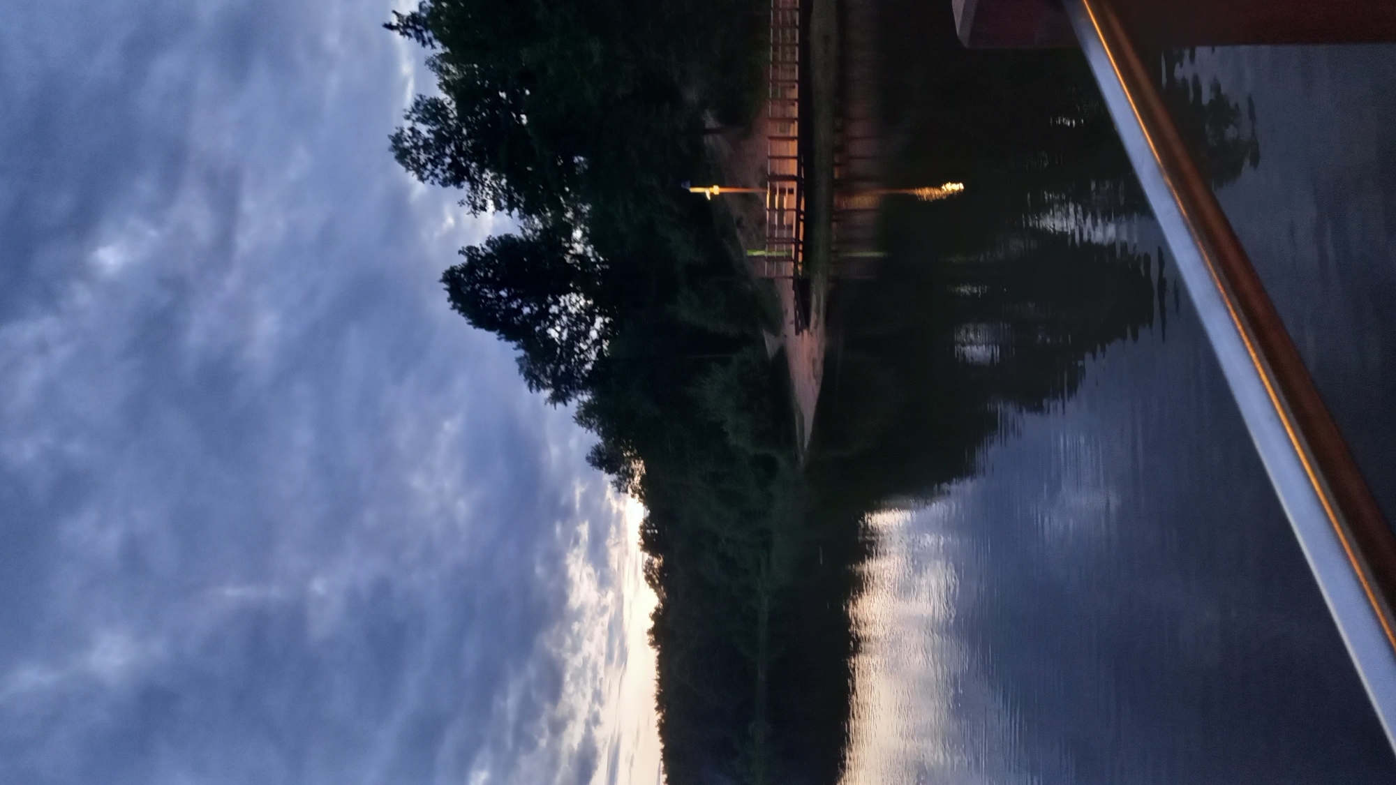 The image shows a waterscape in Krasnogorsk, Russia, likely a lake or reservoir based on the size of the body of water. The lighting suggests it is dusk or twilight. A railing is visible on the right side of the frame, suggesting the photo was taken from a boat or elevated structure. Across the water is a treeline and a pier or walkway with illuminated lights along it. The sky is overcast with dark, heavy clouds. Reflections of the sky and the treeline are visible on the water's surface.