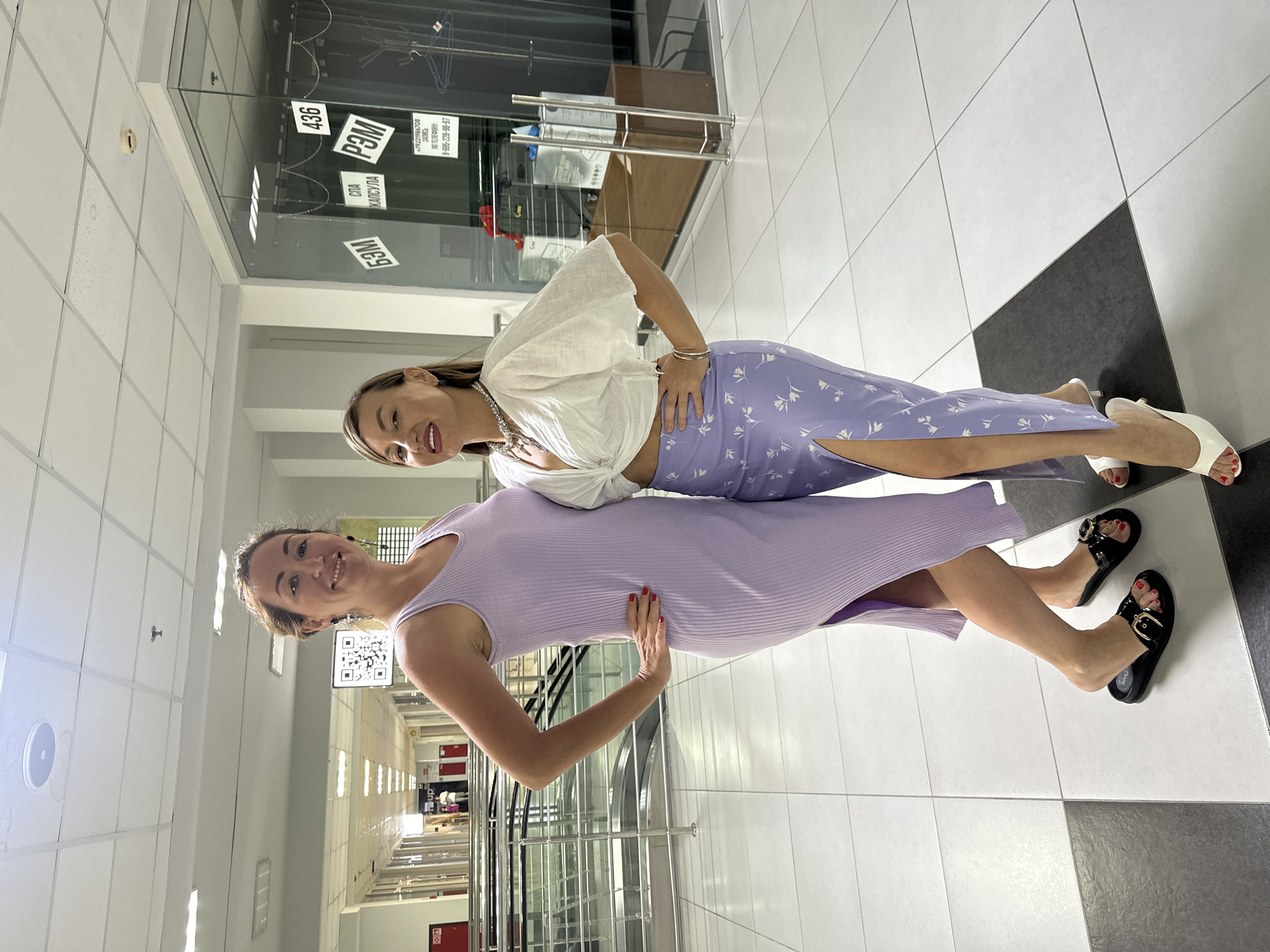 Two women stand facing the camera in what appears to be an indoor public space, possibly a shopping center or a modern office building lobby, with a black and white checkered tile floor. The woman on the left wears a long, light purple ribbed dress with a thigh-high slit and black slides with gold buckles. She has her hands on her hips and is smiling. The woman on the right wears a white cropped blouse with voluminous sleeves and a light purple skirt with a floral pattern and a thigh-high slit. She is also smiling and has her hands on her hips. Behind the women, there is a glass-enclosed area with signs and displays, and in the background, there is an escalator. The lighting suggests it is daytime, and the environment is clean and well-maintained. There is no specific indication that this location is in Sochi, Russia, in the provided image.