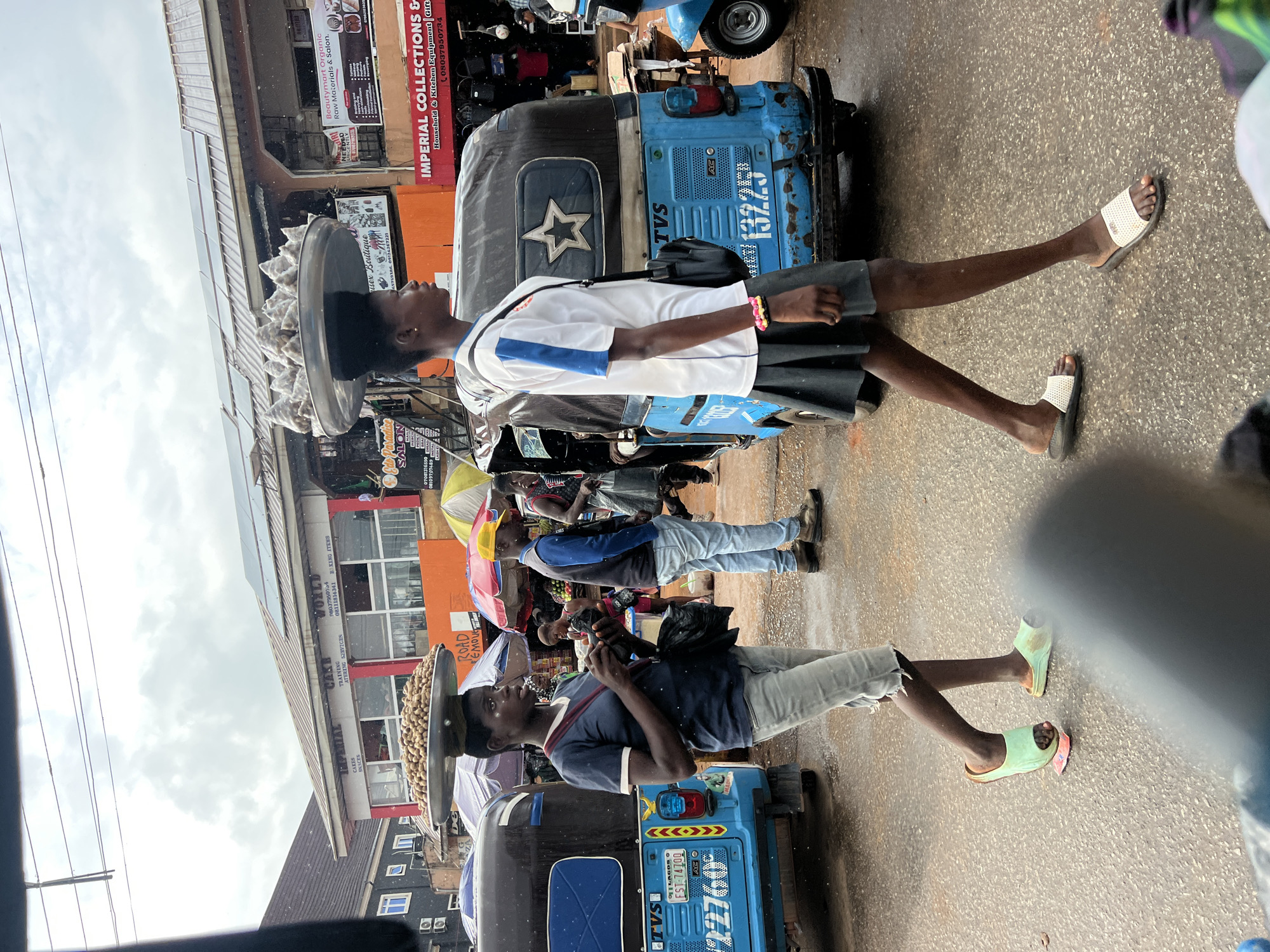 An overcast day in Okpanam, Nigeria, shows two young women carrying goods on their heads as they walk through a bustling street market. The woman on the left, wearing a dark t-shirt and gray shorts, has a large metal tray laden with brown nuts balanced on her head. She carries a black bag on her shoulder and appears to be looking down at something in her hands. She is wearing light green flip-flops. The woman on the right, in a white t-shirt with blue and black trim and a dark skirt, walks with a similar tray of goods, which are conical in shape. She has a black crossbody bag and is wearing white flip-flops with a pink and yellow bracelet on her left wrist. They are walking past blue, three-wheeled vehicles, commonly known as Keke Napep or tricycles, which are prevalent modes of transportation in Nigeria. One of the Keke Napep vehicles has "LAGOS FST-74700" printed on its license plate. In the background, a variety of shops and stalls line the street, with colorful umbrellas and awnings providing shade. The street appears wet, suggesting recent rain. The shops have various signage, including "IMPERIAL COLLECTIONS & HOUSEHOLD & KITCHEN EQUIPMENT" and "Beauty mart Organic Raw Materials & Sales." A man in a yellow cap and blue jacket is also visible walking in the background. The overall scene depicts a vibrant, active marketplace.
